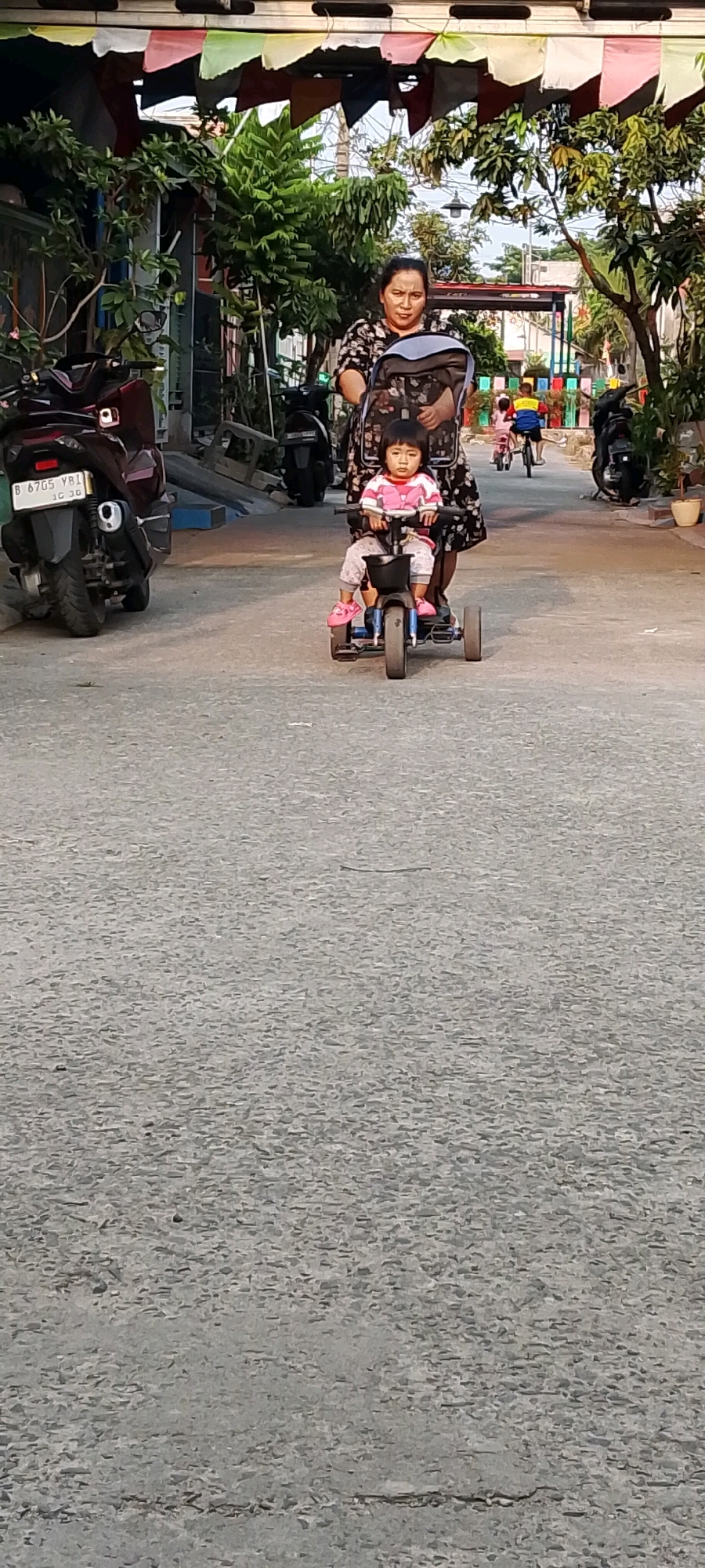 In Satriamekar, Indonesia, a woman pushes a toddler on a tricycle down a paved street. The woman, wearing a dark floral pattern dress, walks behind the tricycle, holding its handle. The toddler, dressed in a pink and white striped shirt and gray pants, sits on the tricycle facing forward, with pink shoes on. The tricycle is blue with a black basket in front.
The street is paved with asphalt and shows signs of wear, with cracks visible. On the left side of the street, a dark red motorcycle is parked. In the background, there are buildings, trees, and colorful banners strung across the street, suggesting a festive or decorated area. Further down the street, another child on a bicycle is visible. The scene appears to be set during daylight hours.