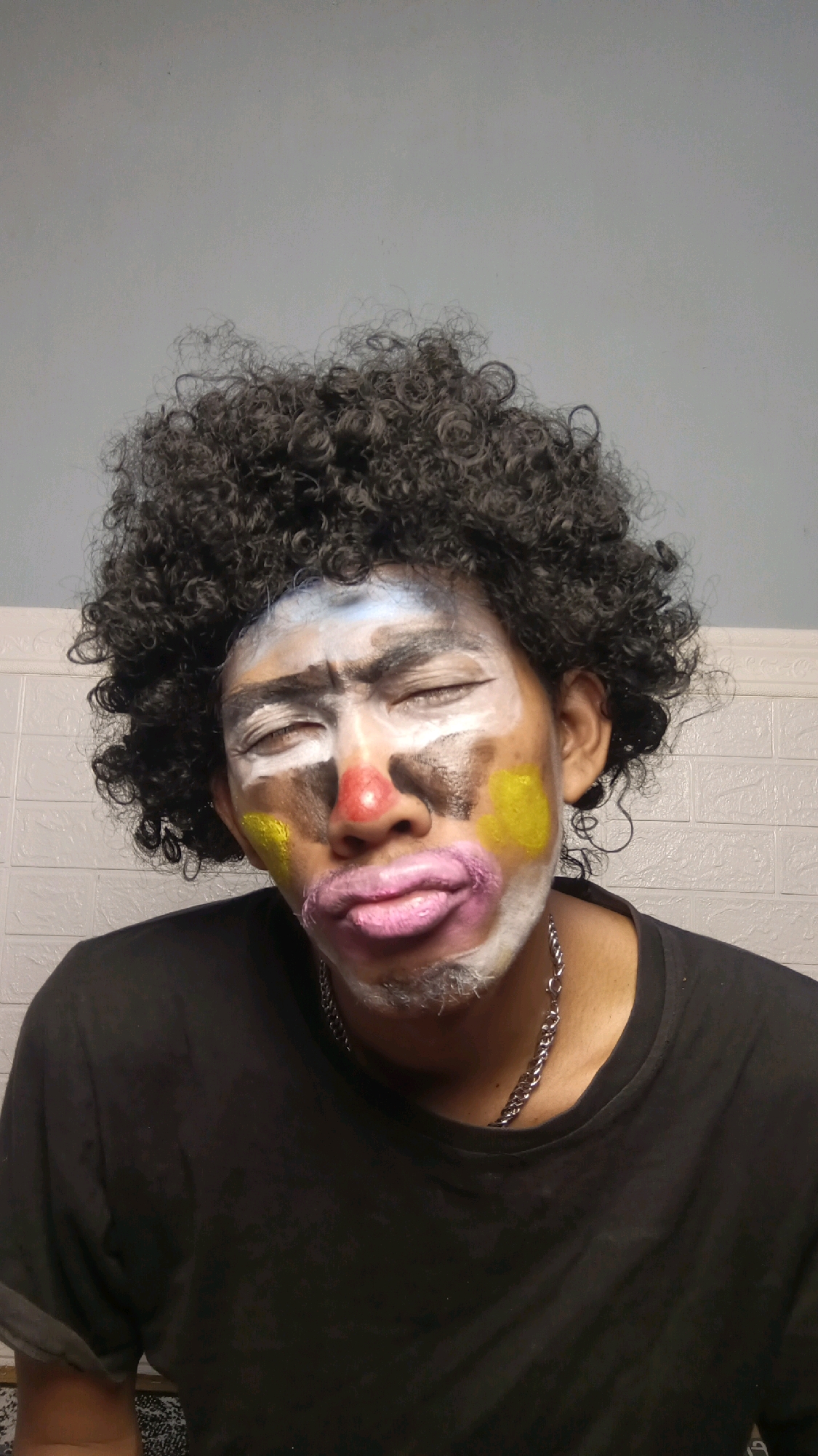 In a portrait taken indoors, likely in Kebumen, Indonesia, a person with dark, curly hair is shown with clown makeup on their face. The makeup includes white and black paint on the face, a red nose, yellow circles on the cheeks, and exaggerated pink lips. The person's eyes are closed. They are wearing a black t-shirt and a silver chain necklace. The background consists of a light-colored wall with a subtle textured pattern.