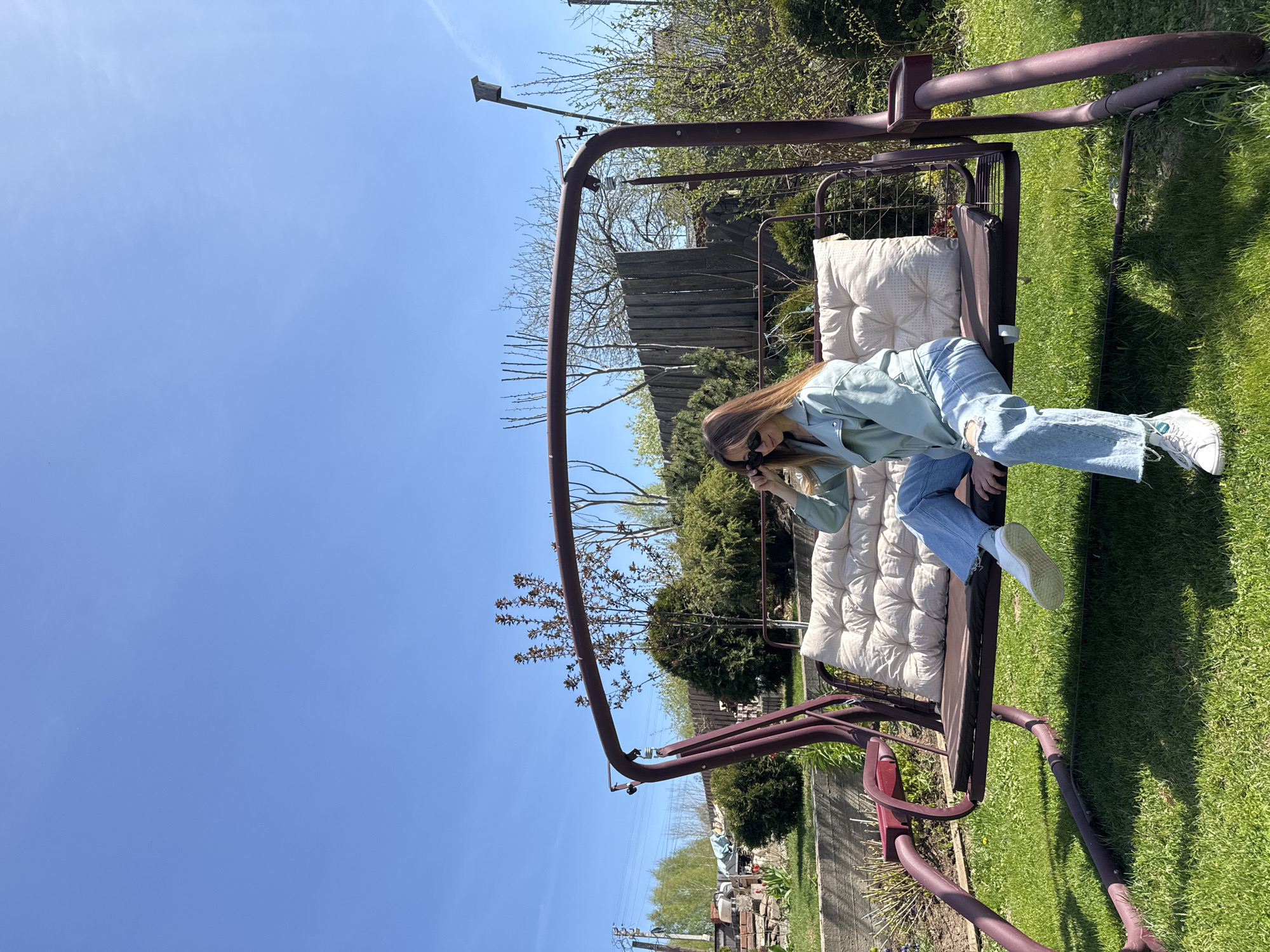 A woman is sitting on a porch swing outdoors, likely in a residential backyard in Kharnavy, Russia. She is wearing a light green jacket, ripped blue jeans, and white sneakers. She has long brown hair and is wearing sunglasses. The porch swing is made of a dark brown metal and has beige cushions. The backyard has green grass, trees, and a wooden fence. The sky is clear and blue. There is a light pole visible in the background.