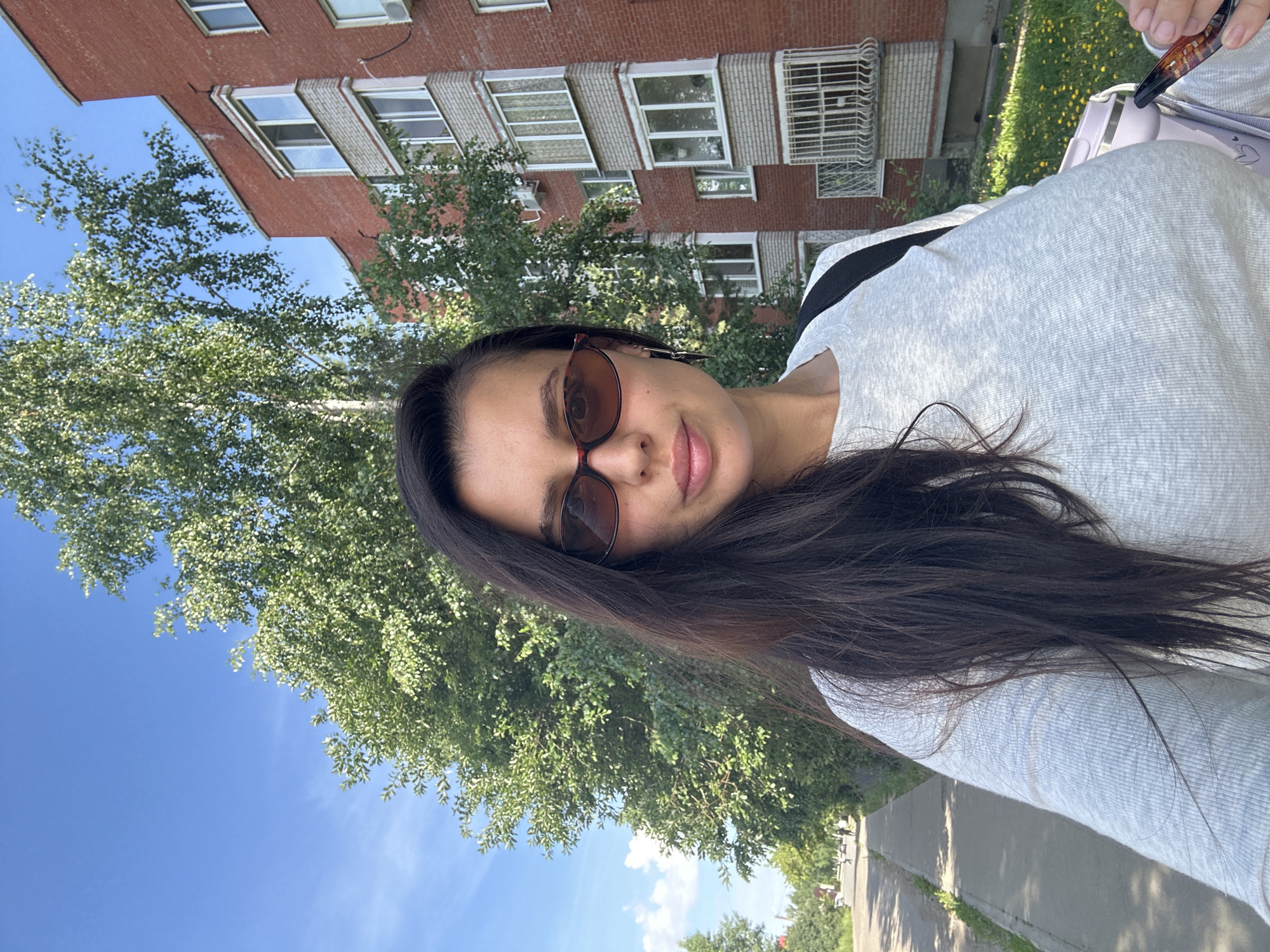 This is a selfie of a young woman taken outdoors. She has long, dark hair and is wearing sunglasses and a light-colored sweatshirt. The woman is smiling slightly, and her face is turned towards the camera. In the background, there is a brick building with several windows, a tall tree with green leaves, and a clear blue sky. To the right of the woman, a street or sidewalk is visible, lined with grass and some small yellow flowers. The image appears to be taken during the daytime, with bright sunlight casting shadows on the ground. The overall mood is casual and relaxed, suggesting the woman is enjoying a pleasant day outdoors in an urban or suburban environment, likely in Chaikovsky, Russia.