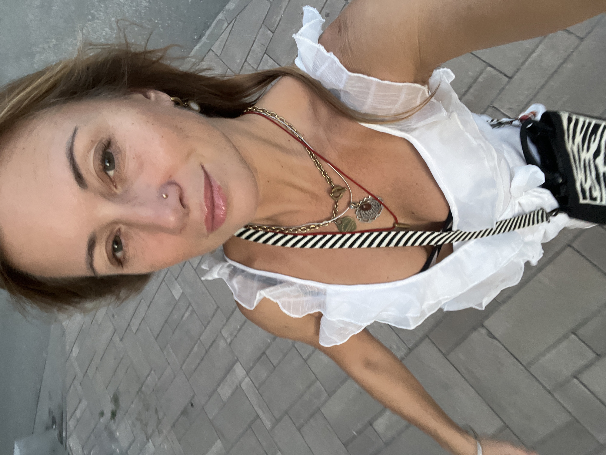 A woman is taking a selfie outdoors on a paved sidewalk. She has blonde hair and is wearing a white ruffled top, a black bra, and multiple necklaces. A black and white striped strap from a bag crosses her chest. The background consists of a textured gray paved surface and a blurred gray wall. The scene appears to be in an urban environment.