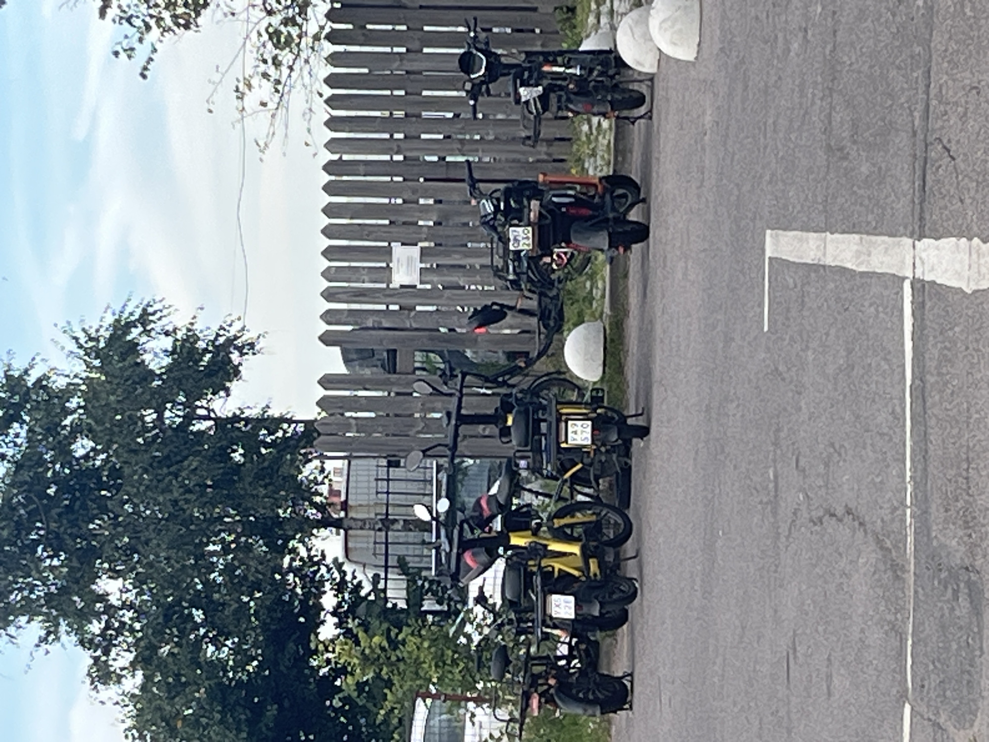 A row of electric scooters and bicycles is parked along a wooden fence in Saint Petersburg, Russia. The scooters and bicycles are predominantly black with some yellow accents. Some of the vehicles have license plates, with visible plates including "YX5 220", "XA9 570", and "QM7 230". White, dome-shaped bollards are placed along the curb in front of the vehicles. The background includes trees, a cloudy sky, and a hint of a building. The scene appears to be an outdoor parking area.