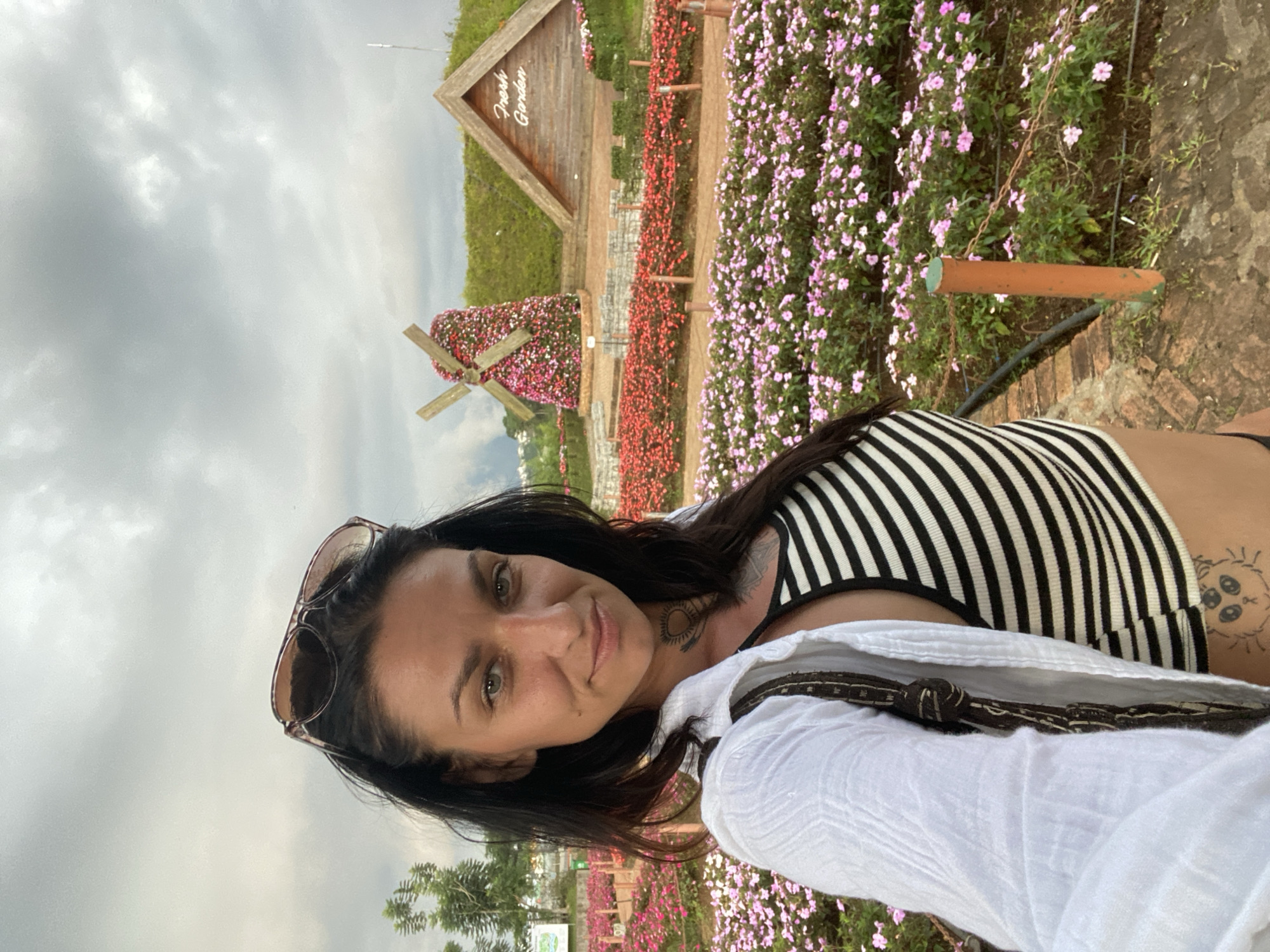 In the city of Cam Ly - Đà Lạt, Vietnam, a woman with dark hair and sunglasses perched on her head poses for a selfie outdoors. She is wearing a black and white striped crop top and an open white shirt. The background features a vibrant flower garden with rows of pink and red flowers, a decorative windmill also covered in flowers, and a wooden structure with the words "Fresh Garden" visible. The sky above is overcast.