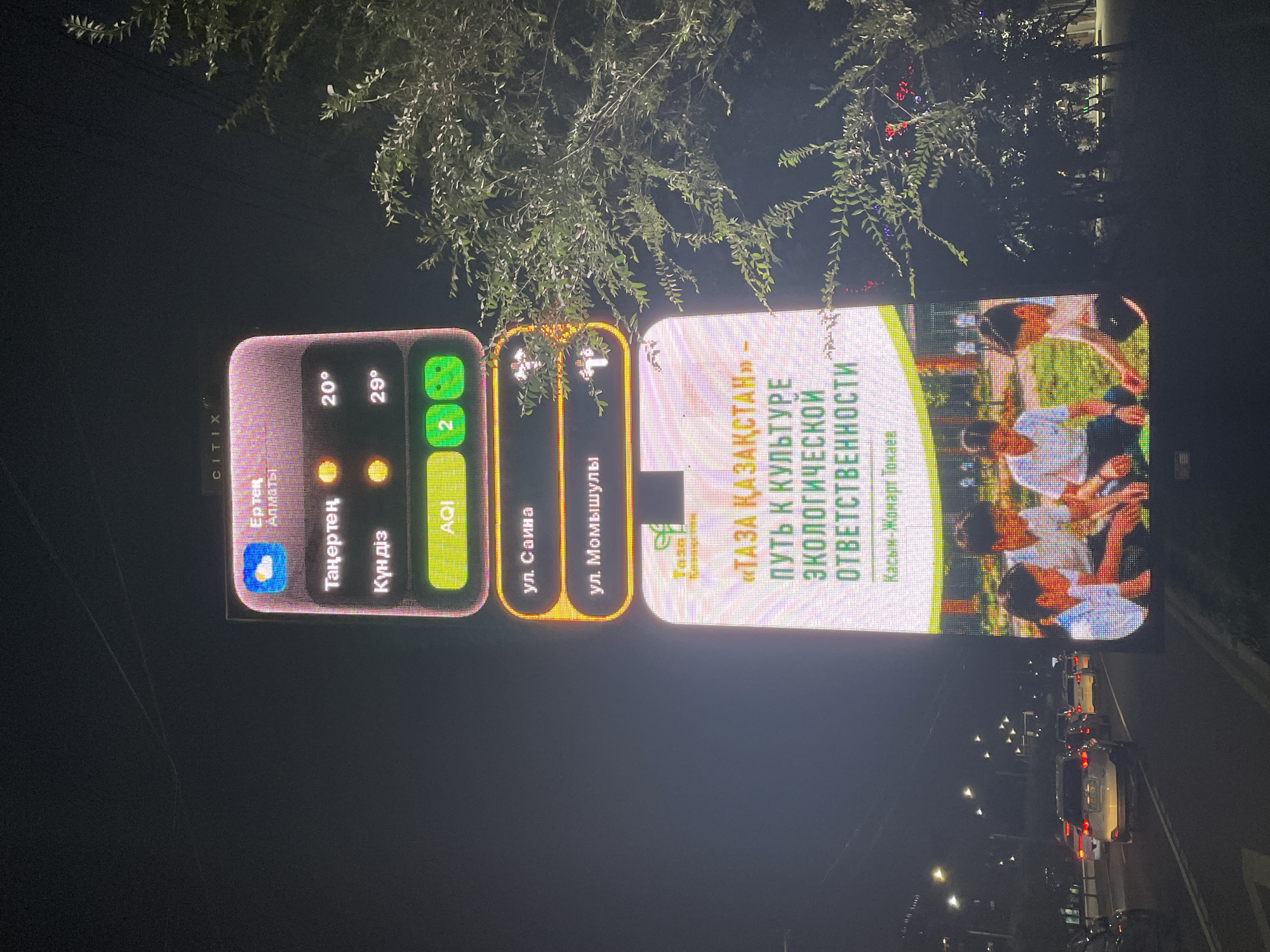 The image shows a large digital billboard at night in Almaty, Kazakhstan. The billboard displays weather information, including the temperature for morning and day, and an Air Quality Index (AQI) reading of 2. It also shows street names, "ул. Саина" and "ул. Мамышулы."
Below this, the billboard features a promotional message in Russian, which translates to "Clean Kazakhstan - the path to a culture of ecological responsibility." It also credits "Kasym-Jomart Tokayev." The bottom part of the billboard shows an image of children sitting on grass in a park-like setting.
The billboard is located in an urban environment, as evidenced by the presence of streetlights and passing cars on a road in the foreground. A tree with leaves is visible on the right side of the image, partially obscuring the billboard. The overall scene depicts a public information display in a city at night.