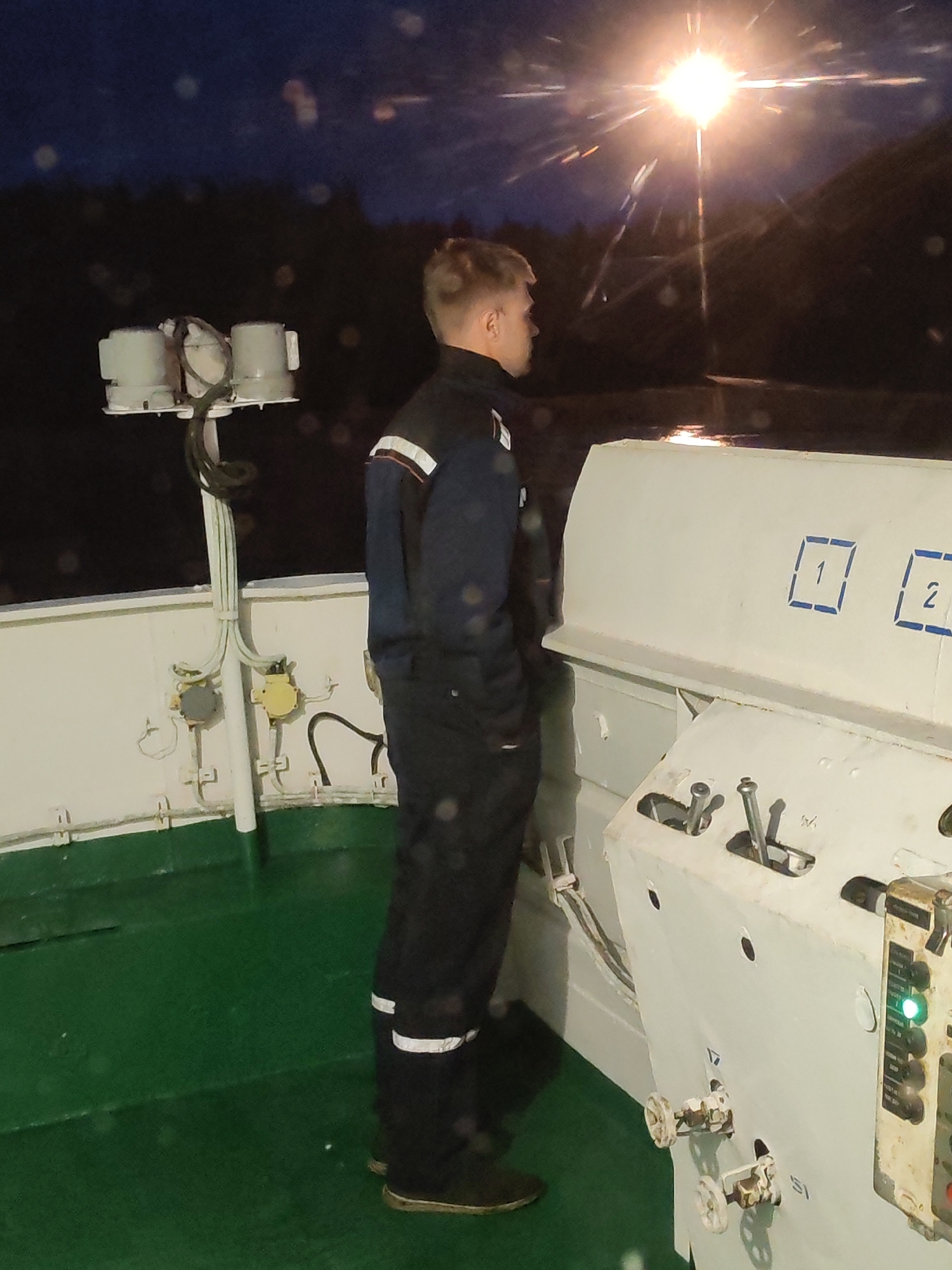 A man stands on what appears to be the bridge of a ship or boat, gazing out at a bright light on the water at night. He is wearing dark work clothes with reflective stripes. The ship's control panel is visible, with levers, dials, and numbered signs. The environment is dark, with the implication of a waterfront setting. There's no specific information to indicate the city of Shalsky, Russia, from the provided image.