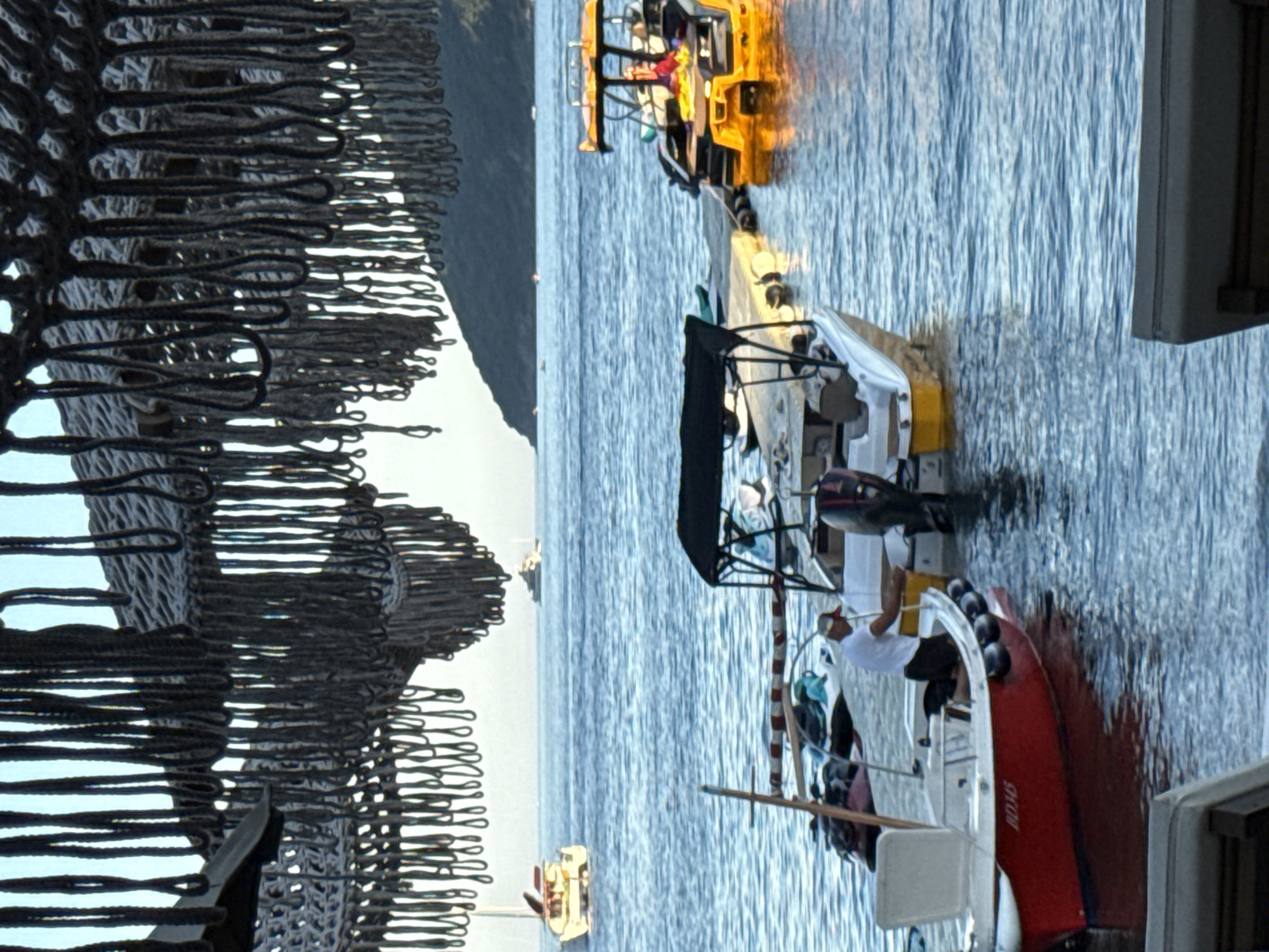 The image captures a sunny day scene on the water near Budva, Montenegro, with several boats docked and at sea. In the foreground, a small red and white boat with the registration number "012345" is moored. A person in a white shirt and black shorts is standing in this boat, attending to something. Next to it, a larger white boat with a black canopy is also docked. Further out, a yellow boat with people on board is tied to a wooden dock, with a jet ski visible nearby. In the background, more boats are present on the calm blue sea, with a hilly coastline visible on the horizon. The scene is framed by a dark, macrame-like structure hanging from above, possibly part of a beachside establishment or shading. The water is clear and blue, reflecting the sunlight.