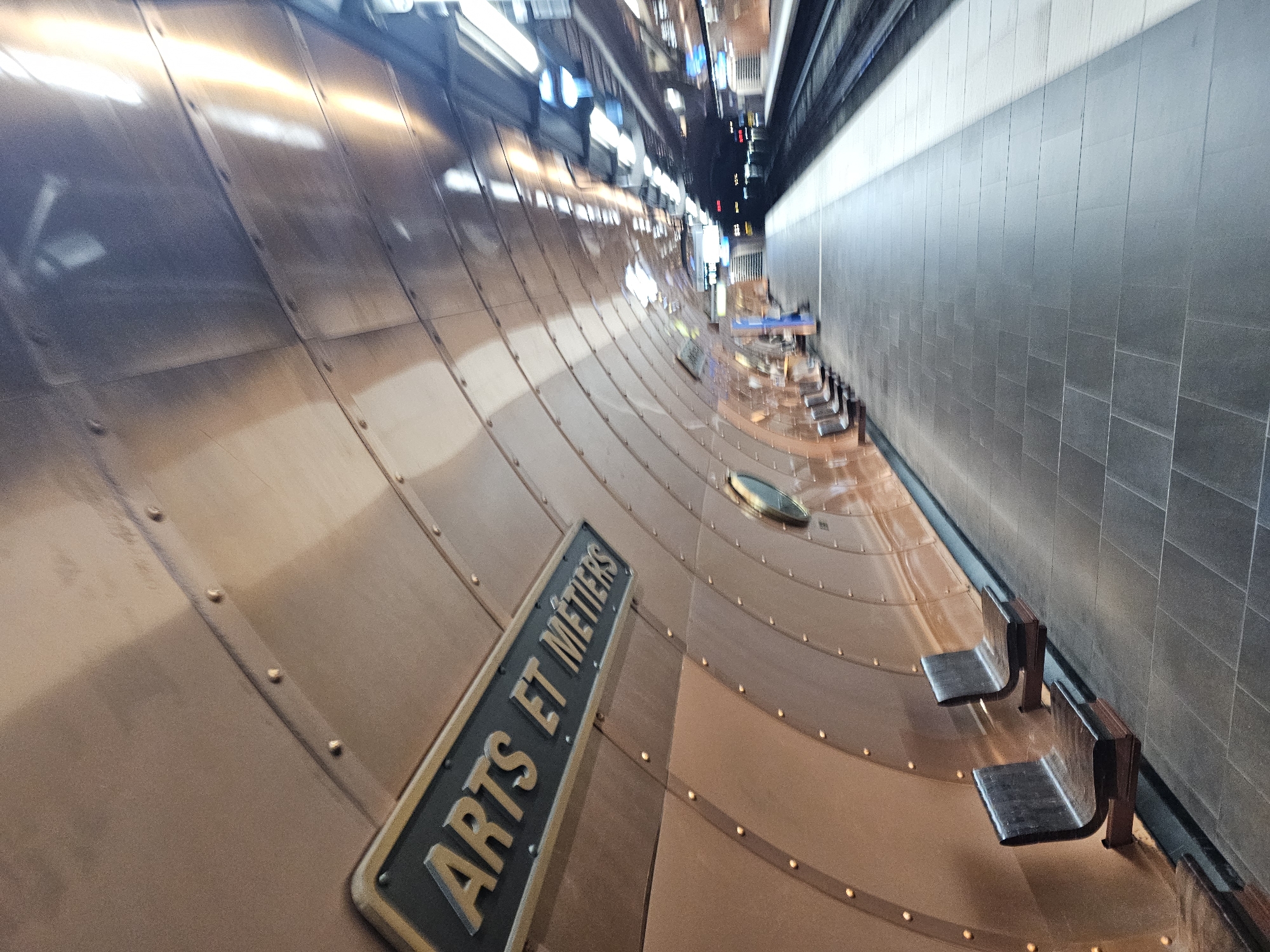 The image shows the interior of the Arts et Métiers metro station in Paris, France. The platform is clad in copper plates riveted together, resembling the inside of a submarine or a Jules Verne novel setting. The station's name is visible on a sign, "ARTS ET MÉTIERS." Metal benches are attached to the copper plating, next to a wall clad in grey tiles. The lighting is bright and reflects off the copper surfaces. The vanishing point is in the distance, where lights and signage can be seen.