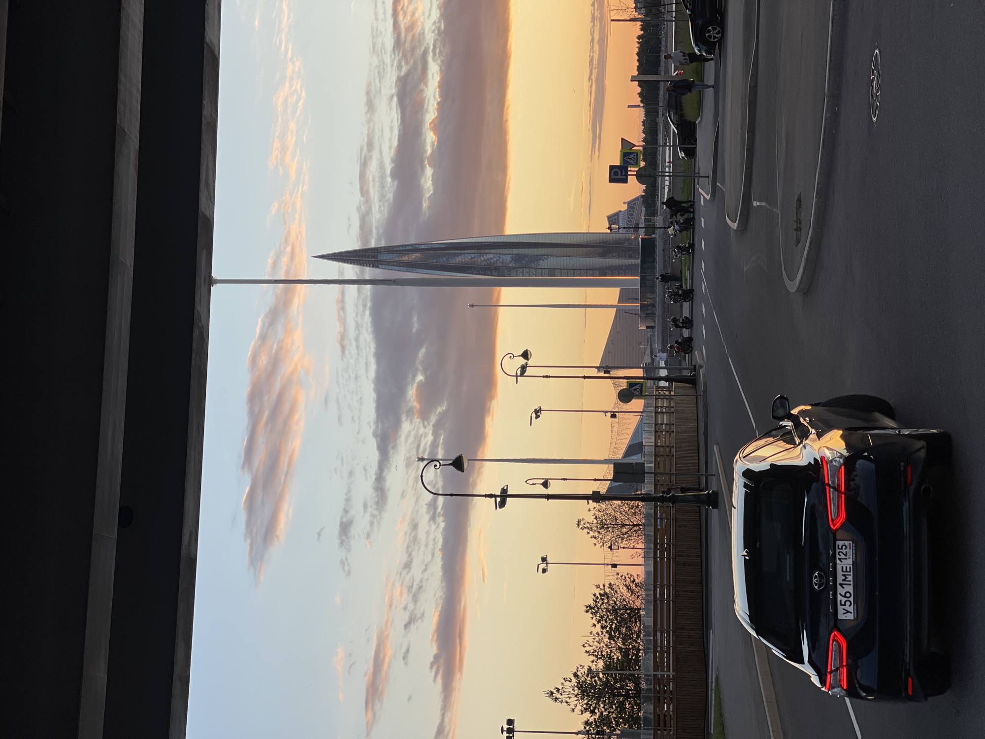 A black Toyota Camry, license plate Y561ME 125, is driving on a paved road under an overpass. The car is the closest subject to the viewer. In the background, a tall, slender, glass skyscraper with a pointed top dominates the skyline, reflecting the warm hues of a sunset. The sky is filled with scattered clouds tinged with orange and pink. Several streetlights with ornate designs line the left side of the road. A group of motorcycles and several pedestrians are visible in the middle ground. The scene is set in a modern urban environment, likely in a city given the presence of the skyscraper and infrastructure. The overall lighting suggests it is late afternoon or early evening.