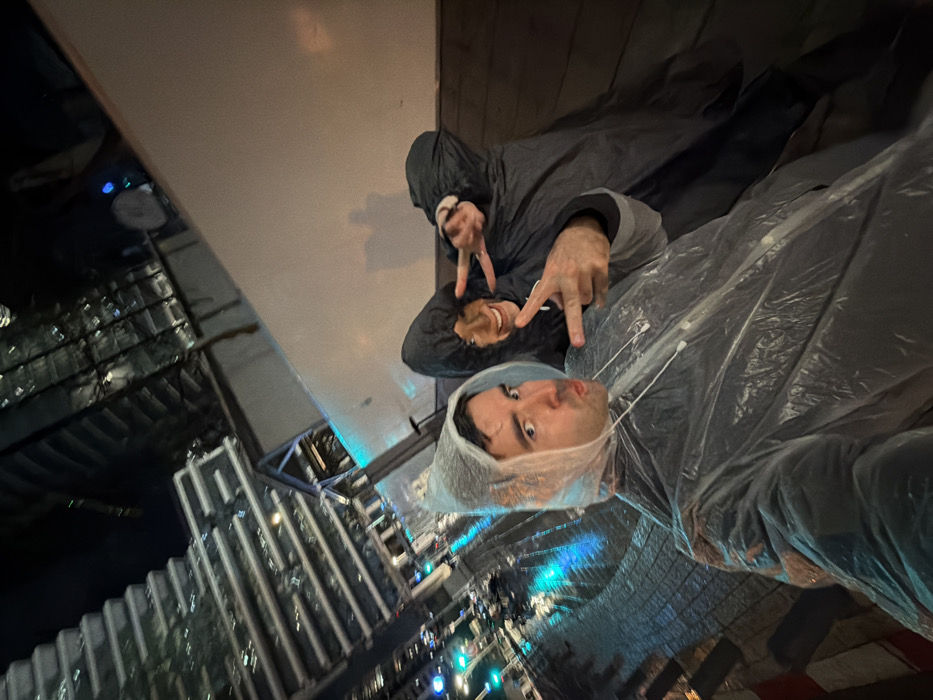 Two people pose in a slightly rotated, nighttime shot. The person in the foreground wears a clear plastic poncho, face visible and mouth puckered. Behind, another person in a darker hooded raincoat makes a peace sign. Tall buildings with lights visible at night are partially seen on the left side of the image.