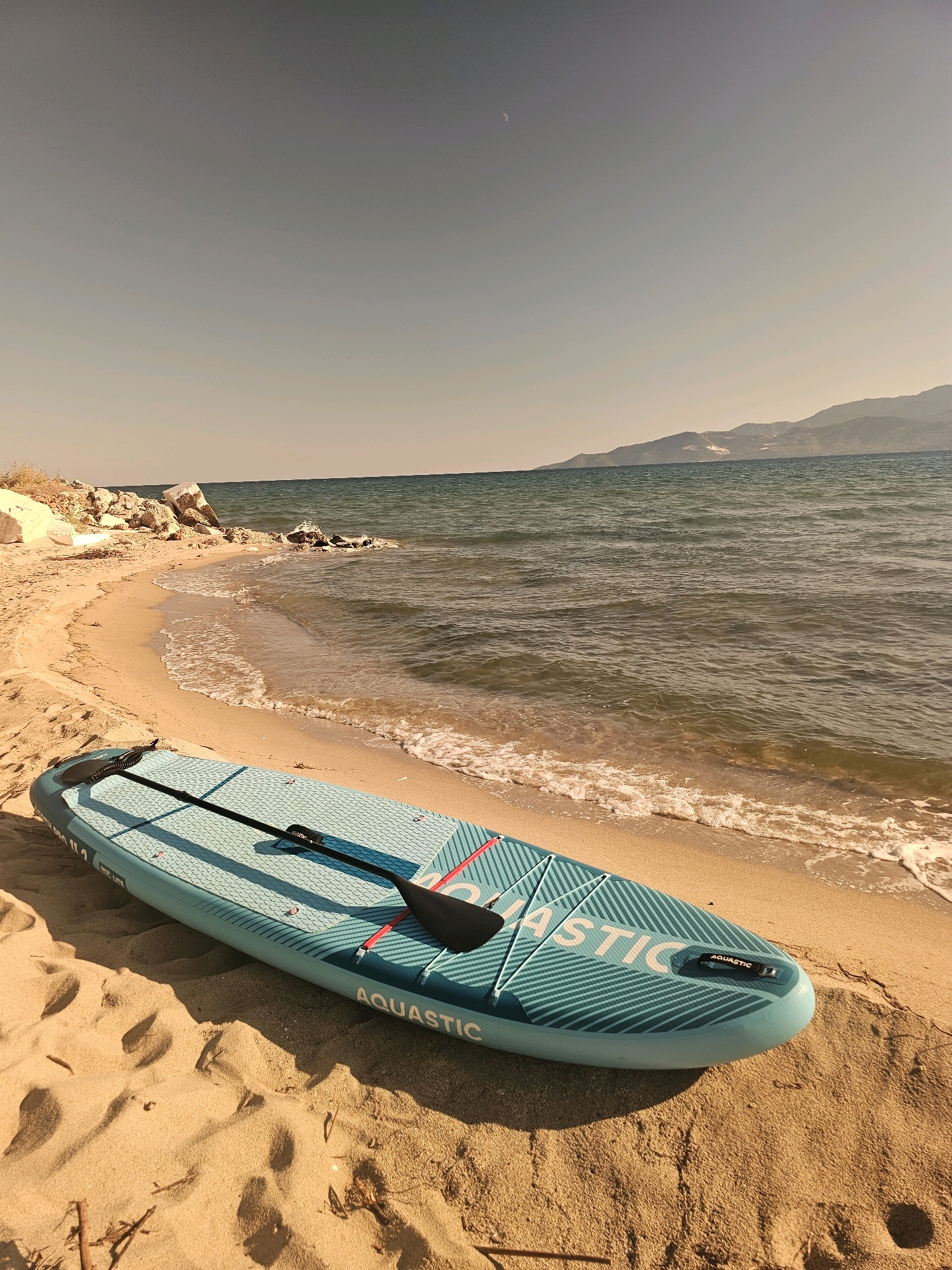 A stand-up paddleboard with a paddle resting on it is lying on a sandy beach. The paddleboard is turquoise and white with "AQUASTIC" written on it. The beach is golden sand and there are small waves gently lapping at the shore. In the background, there is a body of water, a distant coastline with hills, and a clear sky with a crescent moon visible. The overall scene suggests a serene and calm day at the beach, possibly in the early morning or late afternoon due to the lighting.