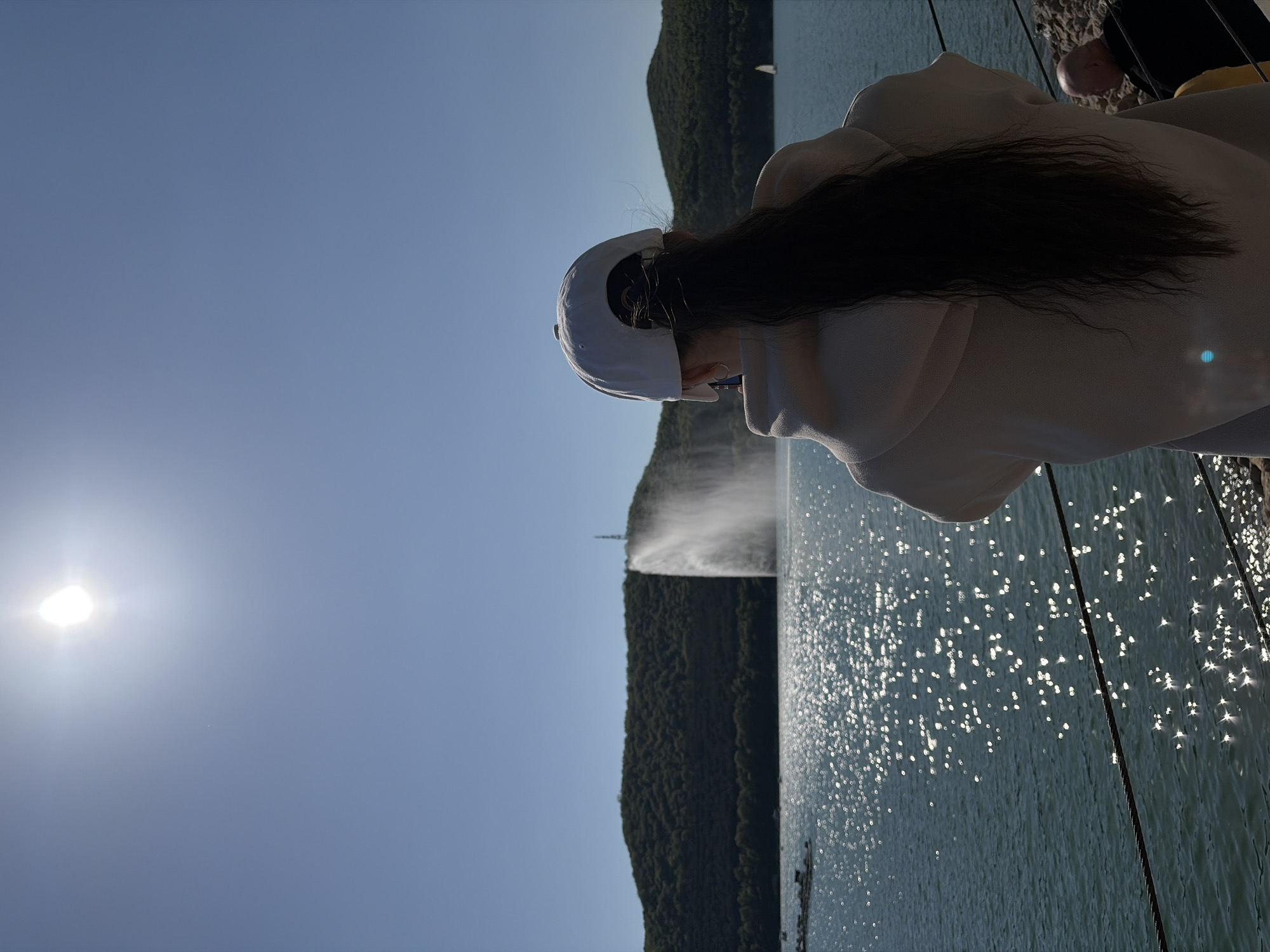 A full shot captures a person wearing a white hooded sweatshirt and a white baseball cap, facing away from the camera, with long dark hair extending down their back. The person is standing beside water, which reflects sunlight. A wooded hill rises from the water’s edge, with a white column of water emanating from near the top of the hill. The sun shines brightly in the sky. This scene is in Abrau-Dyurso, Russia.