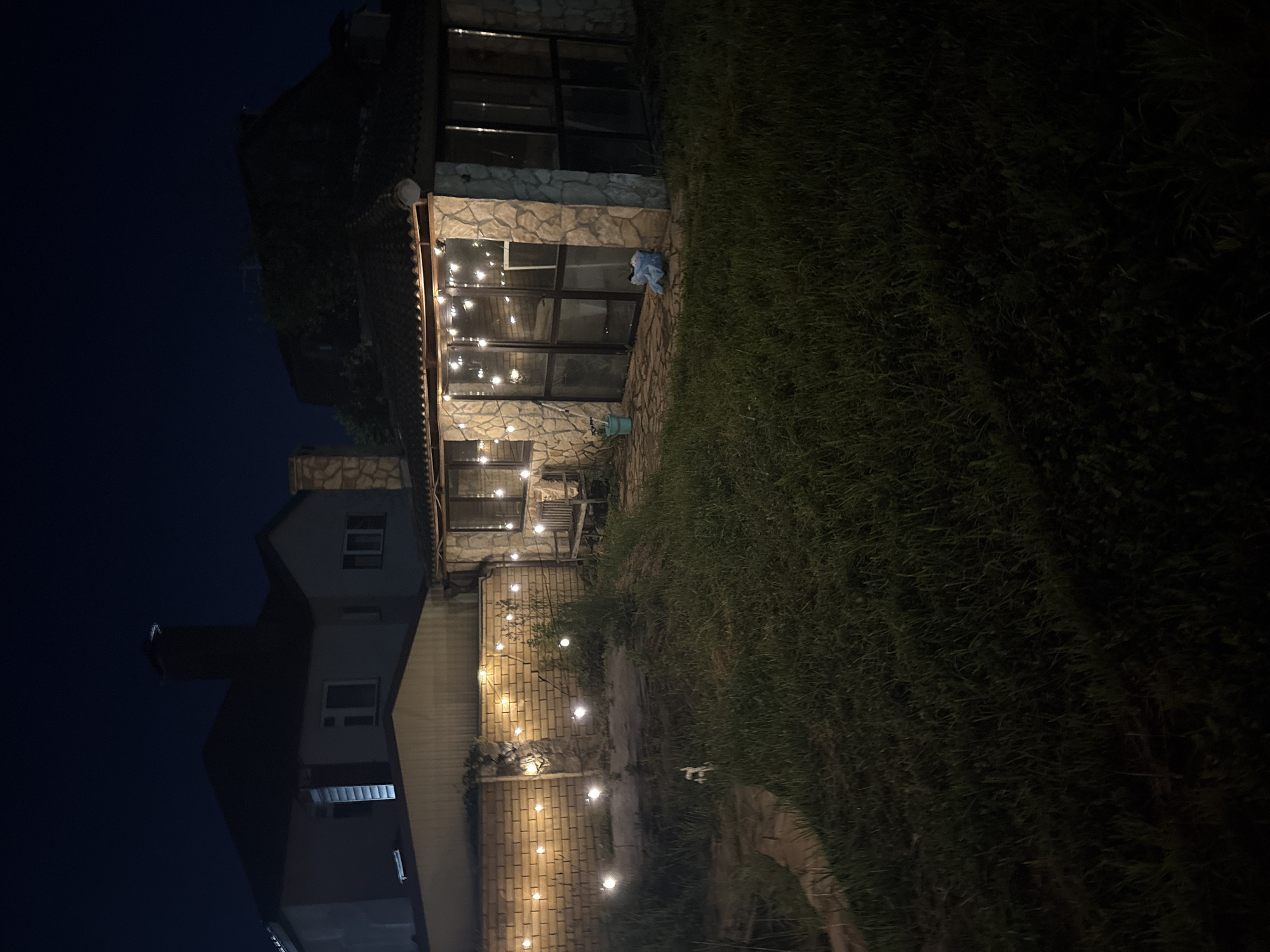 The image depicts a nighttime scene in the city of Orekhovka, Russia, featuring a backyard area adjacent to a two-story house and a stone-clad sunroom or conservatory. The sky is dark blue, indicating it is nighttime.
In the foreground, a grassy lawn covers most of the ground. To the left, a pathway made of irregular stones leads into the yard. A small, light-colored object, possibly a small animal or a white plastic bag, is visible on the grass near the path.
A brick wall runs along the middle ground, decorated with a string of warm white globe lights. The lights are evenly spaced and illuminate the wall and the surrounding area. Beyond the brick wall, a conservatory or sunroom with a tiled roof and stone pillars is visible. This structure has large glass windows and doors, and another string of globe lights is draped along its roofline, casting a warm glow. Inside the conservatory, some furniture and objects can be vaguely seen through the glass. A wooden bench is placed just outside the conservatory.
A larger, darker house is visible in the background to the left, with a prominent chimney. The overall impression is that of a residential property at night, with decorative lighting creating a cozy atmosphere. There are no people visible in the image.