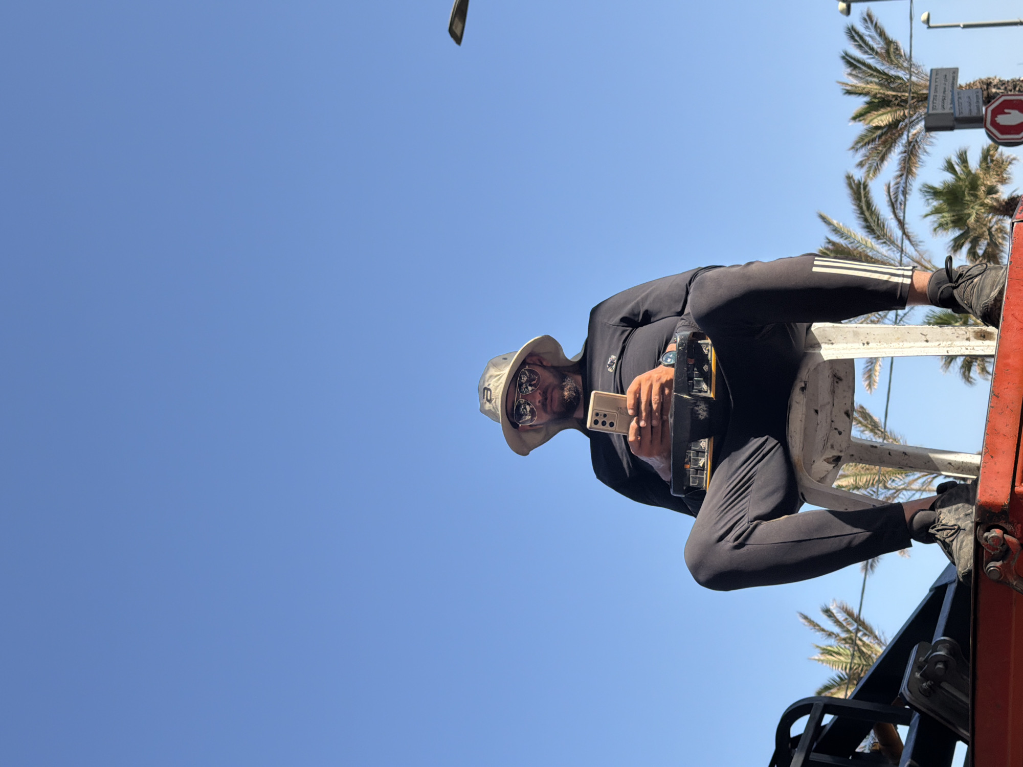 The image, rotated 90 degrees clockwise, presents a dynamic low-angle shot of a man elevated against a vast, clear blue sky. He appears to be seated or perched on a red and white metal platform or lift structure, parts of which are visible in the lower right quadrant and beneath him.
The central subject, a man, is looking down towards the camera, his face partially obscured by dark sunglasses and a light-colored bucket hat. He sports a beard and is dressed in a dark long-sleeved top and dark athletic pants, which display three prominent white stripes on the outer seam of his left leg, reminiscent of Adidas branding. A watch is visible on his left wrist. He holds a light-colored smartphone in his right hand, possibly using it to take a photo or video. His dark sneakers are visible as his feet rest on the platform's framework.
In the upper right portion of the frame, against the clear sky, several palm trees are visible, characteristic of a sunny, coastal environment. Adjacent to the palm trees, parts of a street light arm extend from the top edge. Two street signs are also present: a red circular sign, likely indicating "No Entry" or "Stop," and a rectangular white sign positioned above it. This rectangular sign features text in both Arabic and Hebrew. The Arabic text "لا لانتظار المركبات" (No waiting vehicles) is partially visible, indicating a "no parking/no waiting" regulation, further reinforced by similar Hebrew text below it.
The brilliant sunlight and unblemished blue sky suggest a bright, clear day, likely midday or afternoon. The combination of palm trees, modern urban infrastructure, and multilingual street signs strongly points to an urban setting in Tel-Aviv, Israel, as per the context. The scene captures an individual engaged with his phone from an unusual elevated vantage point.