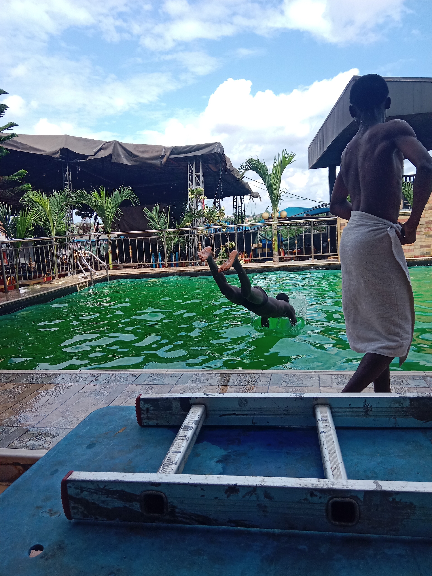 In Nkpor, Nigeria, on a clear day with scattered clouds, a young Black male is captured mid-dive into a vibrant green swimming pool. His body is arched, legs extended, and arms reaching forward as he breaks the water's surface, creating a splash. Another young Black male stands on the poolside, facing away from the camera and towards the diving individual. He is wearing a white towel wrapped around his waist. The pool area is bordered by a tiled walkway and a metal railing. In the foreground, a blue table holds a silver ladder. Behind the pool, a dark tented structure, palm trees, and buildings are visible, suggesting an outdoor event or entertainment venue. The scene captures a moment of recreation and leisure.