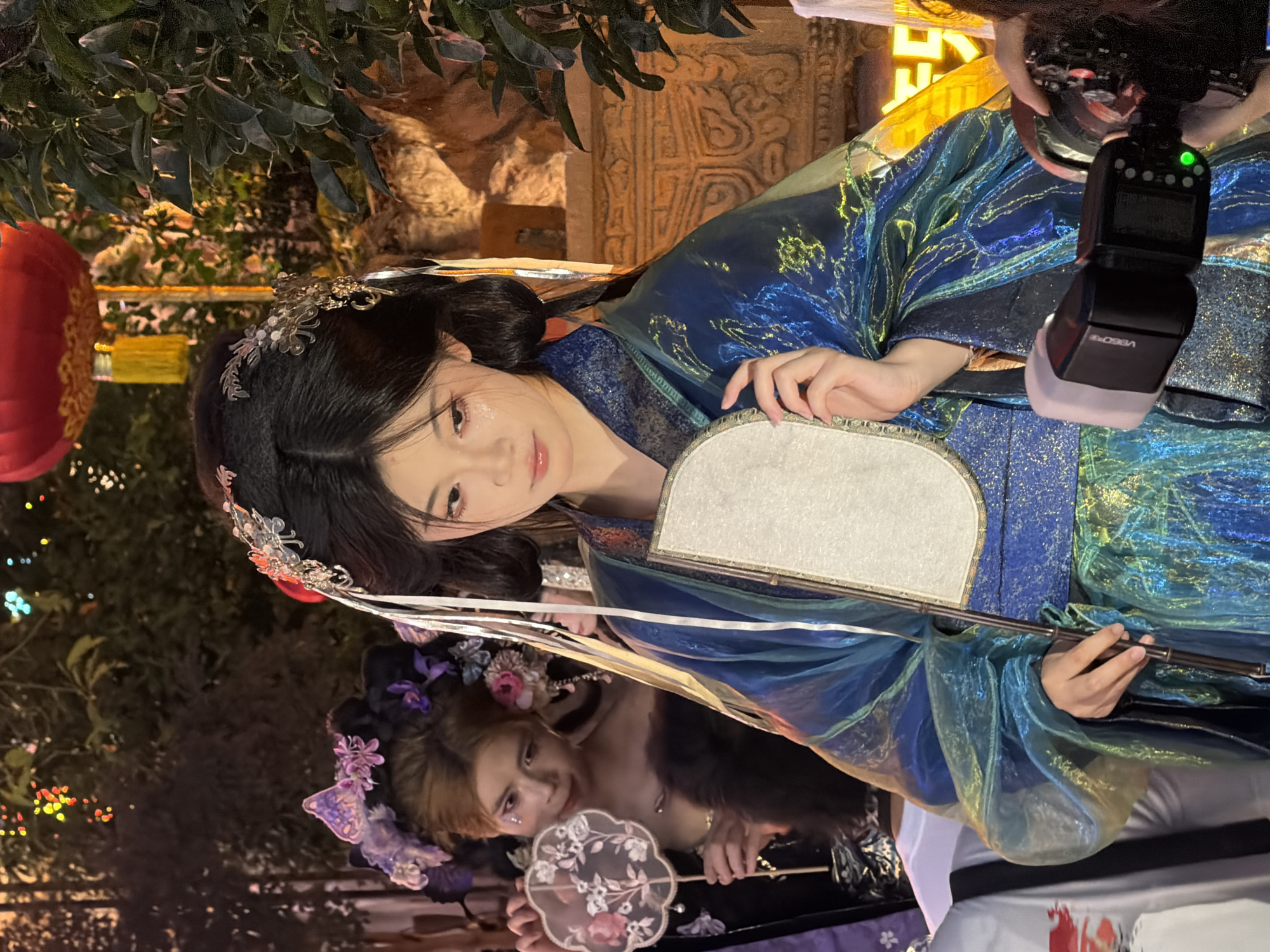 In an outdoor setting at night, possibly a street or courtyard in Yuzhong District, China, illuminated by festive lights and a red lantern, a young East Asian woman stands in the foreground, adorned in a blue and green iridescent traditional Chinese garment with intricate embroidery. She wears an elaborate headdress with delicate floral and metallic accents. In her right hand, she holds a fan made of white material with a floral design. Her makeup includes glitter on her cheeks and eyelids.
Behind her and slightly to the left, another East Asian person, also dressed in elaborate traditional attire with a floral headdress, holds a similarly designed fan. The background features foliage and what appears to be a stone structure with decorative carvings. A camera flash unit is visible in the lower right corner of the frame, suggesting the image was taken during a photographic event or photoshoot. The overall ambiance is celebratory and artistic, evoking a sense of traditional Chinese culture.