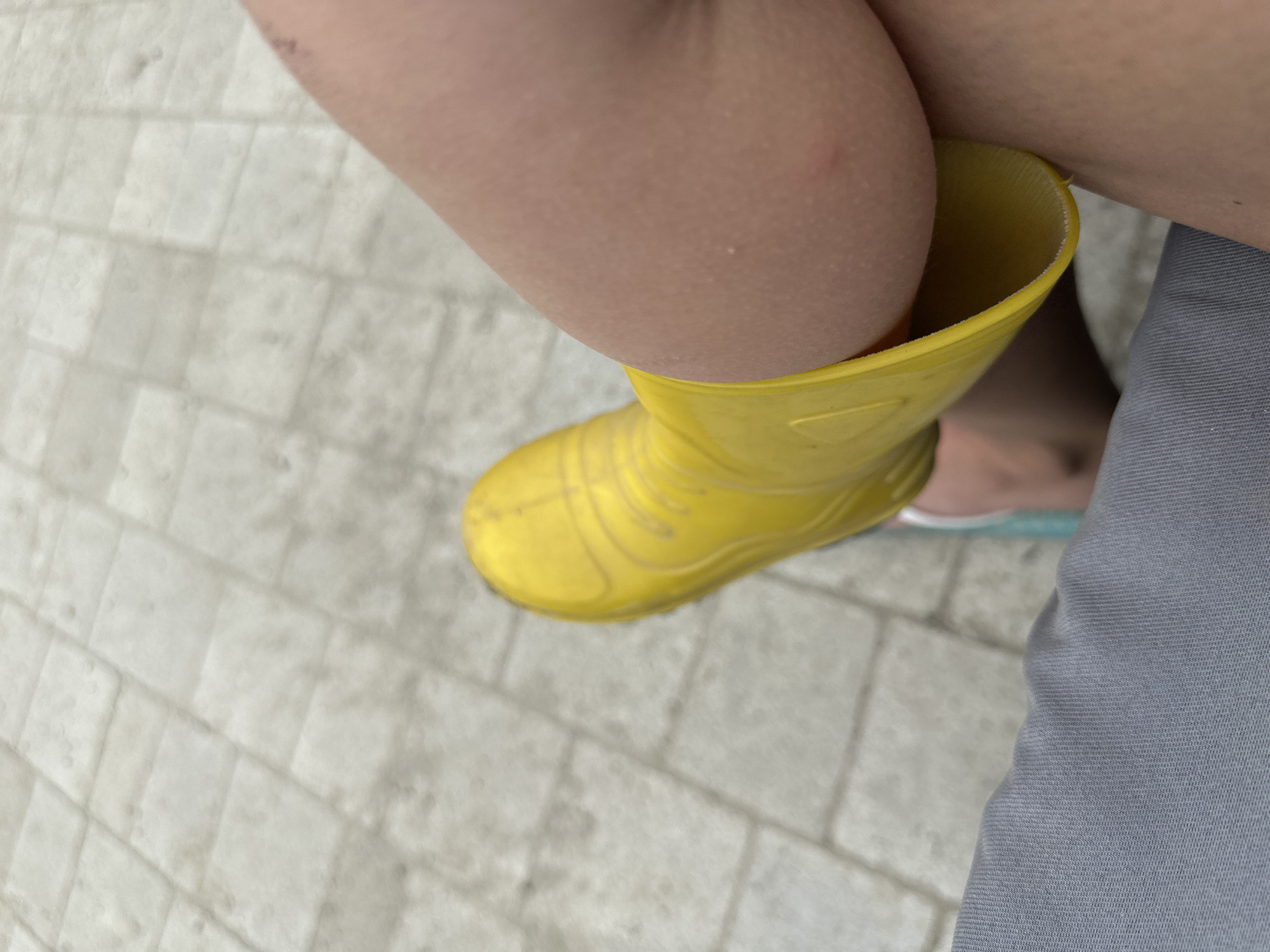 The image shows a close-up, high-angle view of a person's legs. One leg is encased in a yellow rubber boot, with the opening of the boot resting against the other leg. The person is wearing gray pants on the lower portion of their body. The ground beneath them appears to be a paved surface made of light-colored rectangular tiles, suggesting an outdoor urban setting. The scene does not contain any specific details that would allow for identification of the location as Malinovka, Russia.