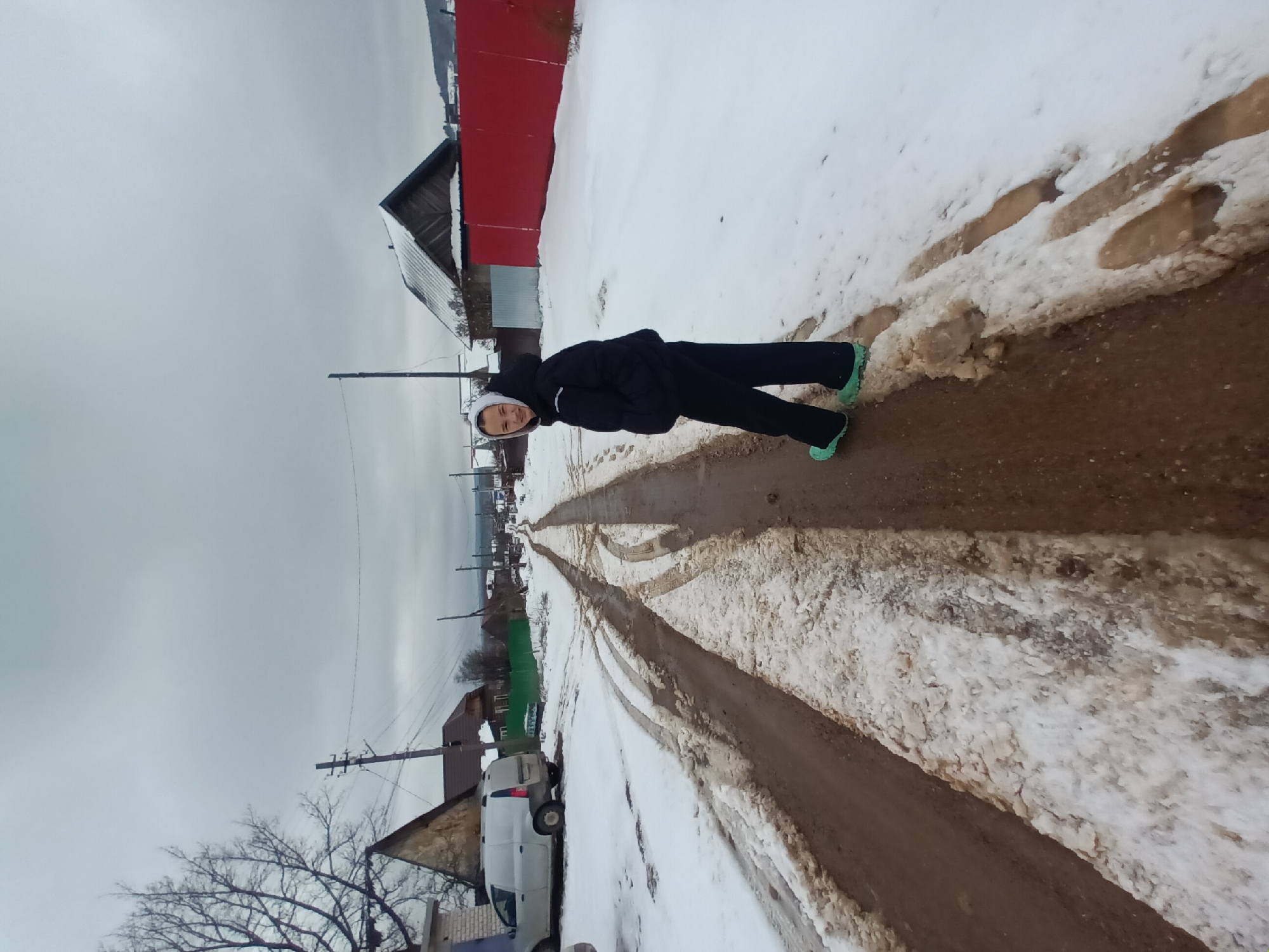 The image shows a person lying on their side on a muddy road, likely a village street. The scene is in Ольховка, Russia. Patches of snow are visible on both sides of the road, which has two tracks running down it. The person is wearing dark clothing and light-colored shoes. Behind the person are several houses, some with red or green paint. A white van is parked near one of the houses. Utility poles and bare trees line the street. The sky is overcast.