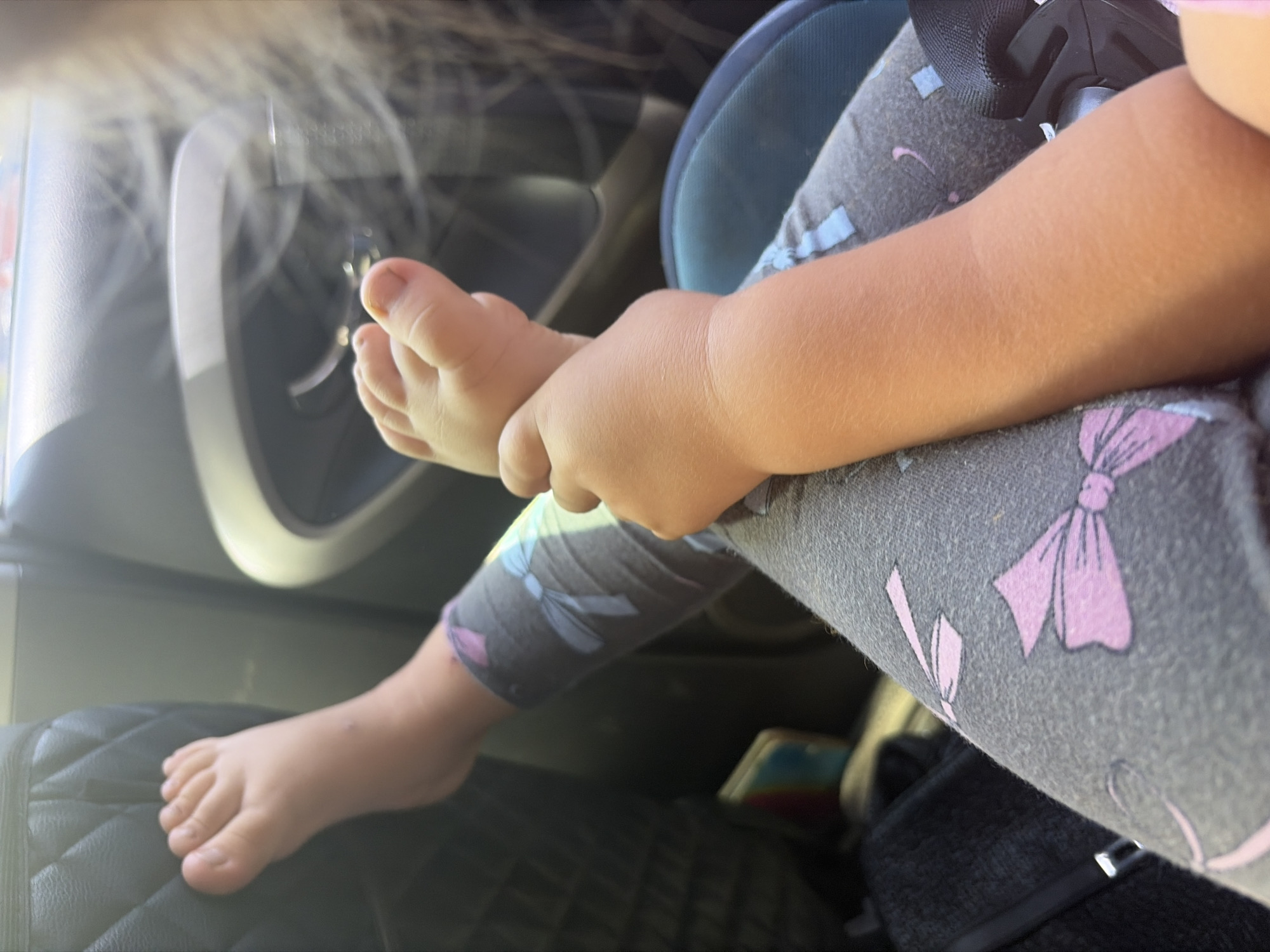 In the interior of a vehicle, a close-up shot focuses on the legs and feet of two young children. One child's bare foot, with visible toes, rests against the dark, quilted upholstery of a car seat. This foot is positioned above the other child's legs, which are covered in grey leggings adorned with pink bow patterns. The second child's arm is visible, holding onto the first child's foot. The background suggests the car's interior, with a car door handle visible and a hint of sunlight filtering in. The scene appears to be captured during daytime travel, but no specific location in Rostov-on-Don, Russia, can be determined from the visual information provided.