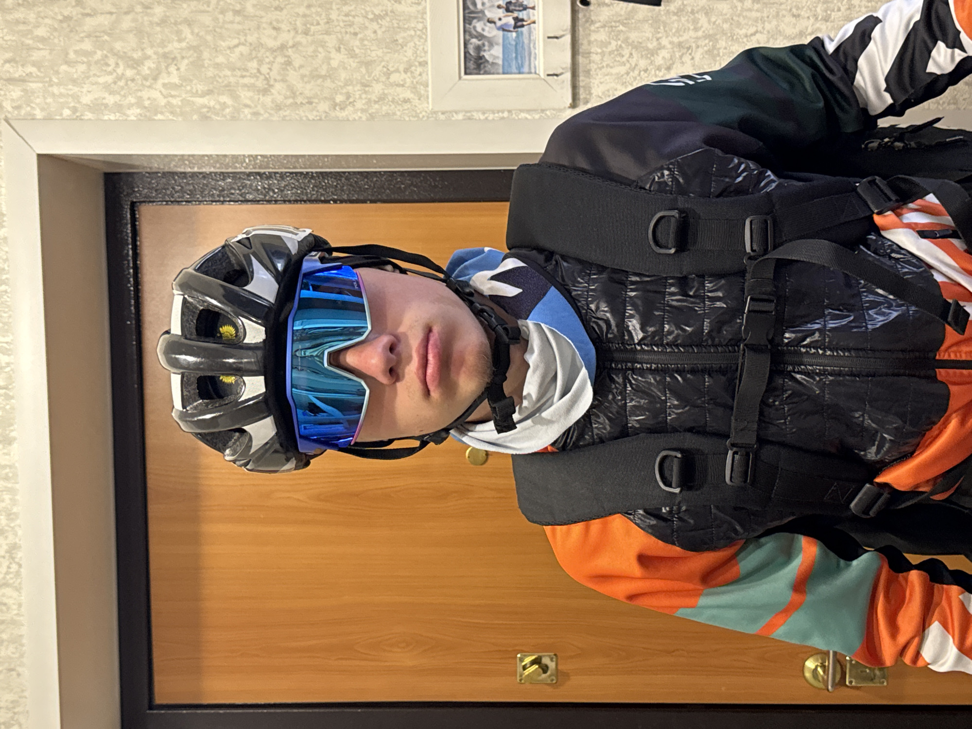 The image depicts a person wearing a cycling helmet and sunglasses. The person is also wearing a black quilted vest with orange and green sleeves, and a backpack. The background shows a light-colored wall, a picture frame, and a wooden door with a black frame. The scene appears to be indoors, possibly in a home or apartment. The location cannot be determined as Chaikovsky, Russia based solely on the visual elements of the image.