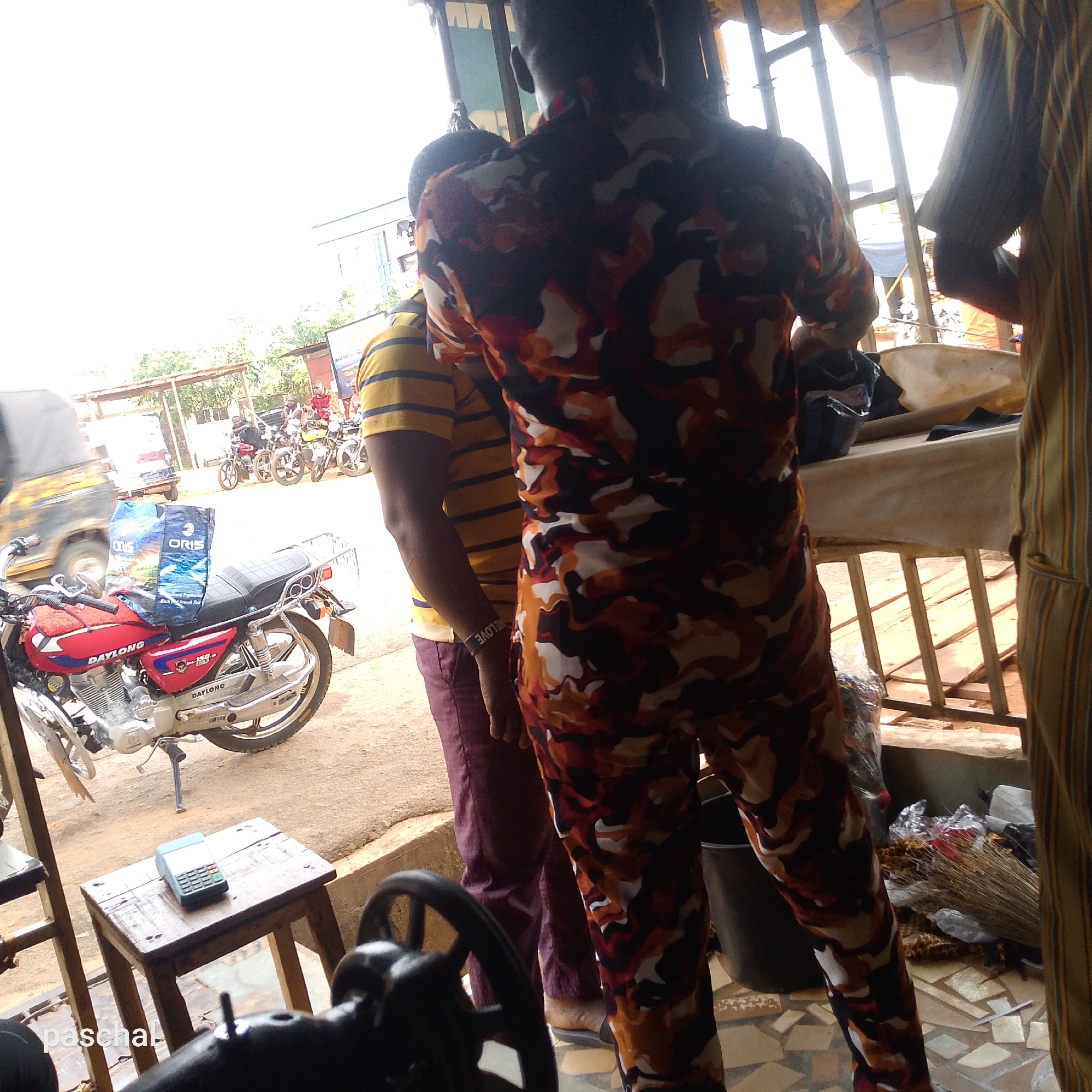 Captured from inside a building in an unidentifiable city, the photograph presents a scene of daily life. A red Daylong motorcycle is parked just outside, with a bag labeled "ORIS" resting on its seat. Several other motorcycles and vehicles are visible in the background, indicating an urban environment.
Inside the building, a sewing machine occupies the foreground, resting on a small wooden stool alongside a calculator. Three people are partially visible, their attire and positioning suggesting interaction or transaction. One person wears a brown, orange, and white camouflage-patterned outfit, another is dressed in a yellow and blue striped shirt and red pants, and a third is wearing a striped yellow and tan shirt. The floor of the building is covered in small, square off-white tiles. A broom and various items are scattered around the base of the frame.