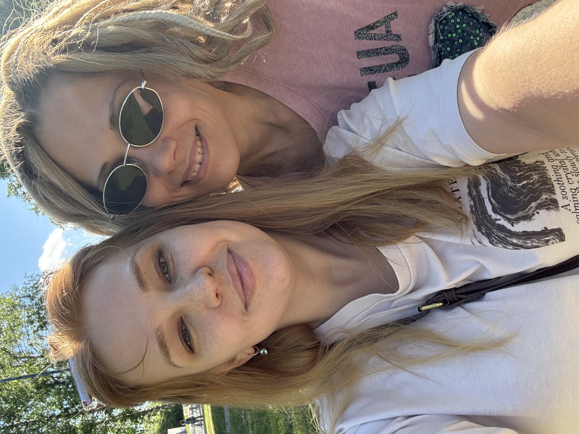 Two women are posing for a selfie outdoors. The woman in the foreground has light red hair, fair skin with freckles, and is wearing a white T-shirt with a dark design on it, and a dark strap over her shoulder. The woman in the background has blonde dreadlocks, is wearing round sunglasses, and a pink t-shirt. They appear to be in a park-like setting with trees and grass visible in the background, possibly in the city of Rusanovo, Russia. The sky is visible in the upper left corner.