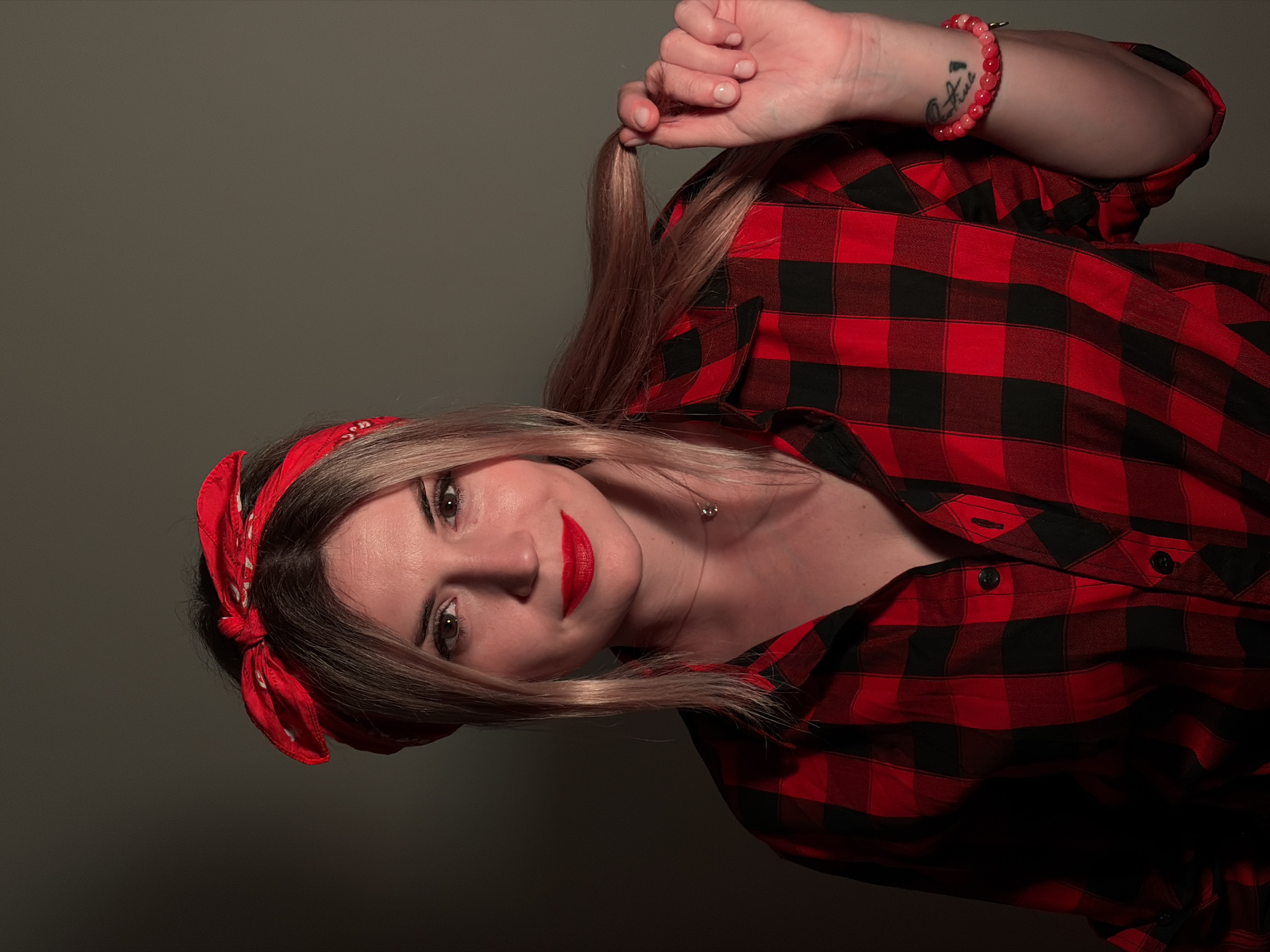 A fair-skinned woman with shoulder-length blonde hair styled with a red bandana is photographed indoors against a dark gray background. She wears a red and black plaid shirt, unbuttoned at the top. Her makeup includes red lipstick, and she wears a thin necklace. She holds a strand of her hair in her hand, and a beaded bracelet is visible on her wrist, which also has a tattoo. The image is rotated slightly clockwise. There is no information about the location in the city of Budva, Montenegro.