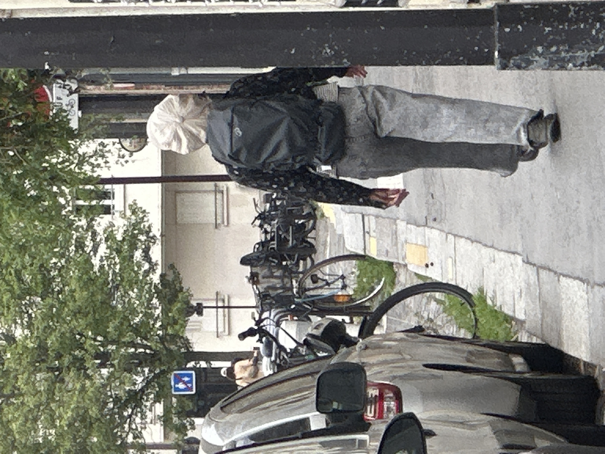 The image shows a person walking on a sidewalk in Paris, France. The person is wearing light-colored pants and a patterned top, a white covering on their head, and a dark backpack. They are walking away from the camera. Behind the person are parked bicycles. A silver vehicle is partially visible at the bottom of the image. A tree is on the left side of the image, and a blue sign is visible in the background.