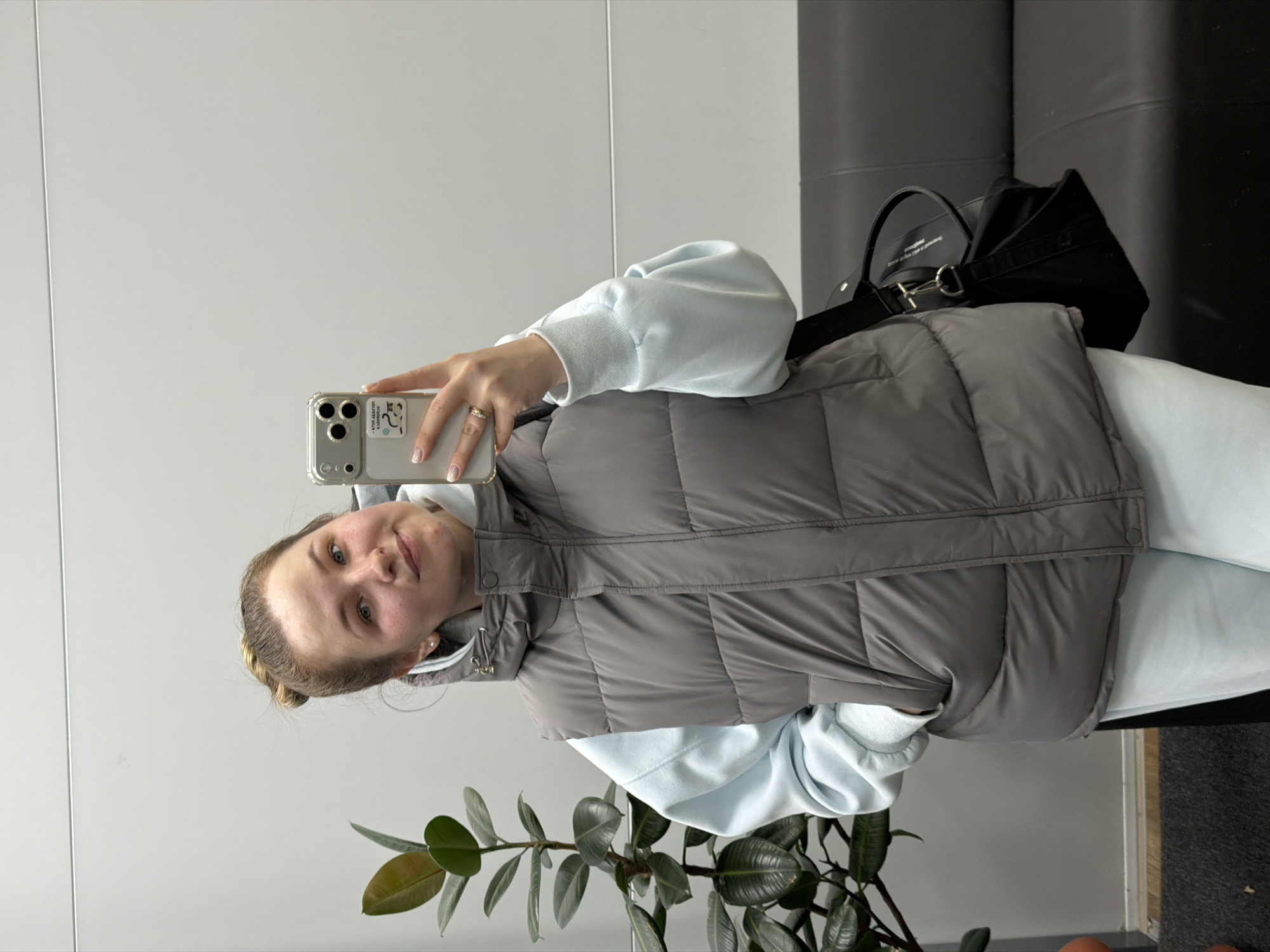 A young woman takes a mirror selfie in an indoor setting. She appears to be in her 20s or early 30s, looking directly at the camera with a soft, pleasant smile. Her hair is pulled back, and she wears a light blue long-sleeved top, possibly a hoodie, underneath a grey puffer vest. Light-colored pants, likely sweatpants, are visible.
She holds a smartphone with a clear case in her right hand to capture the reflection. A ring is visible on her right hand. On the back of the phone case, there's a sticker with partially visible text, including fragments like "ATOS" and "COSMOS" or "LOVELY."
The background consists of plain, light-colored walls. Behind her, a dark piece of furniture, possibly a couch or bench, holds a black tote bag with unreadable text on it. In the bottom left foreground, a potted plant with large green and reddish-brown leaves adds a touch of nature to the minimalist interior. The overall impression is a casual moment in a clean, modern space. The time of day is not determinable.