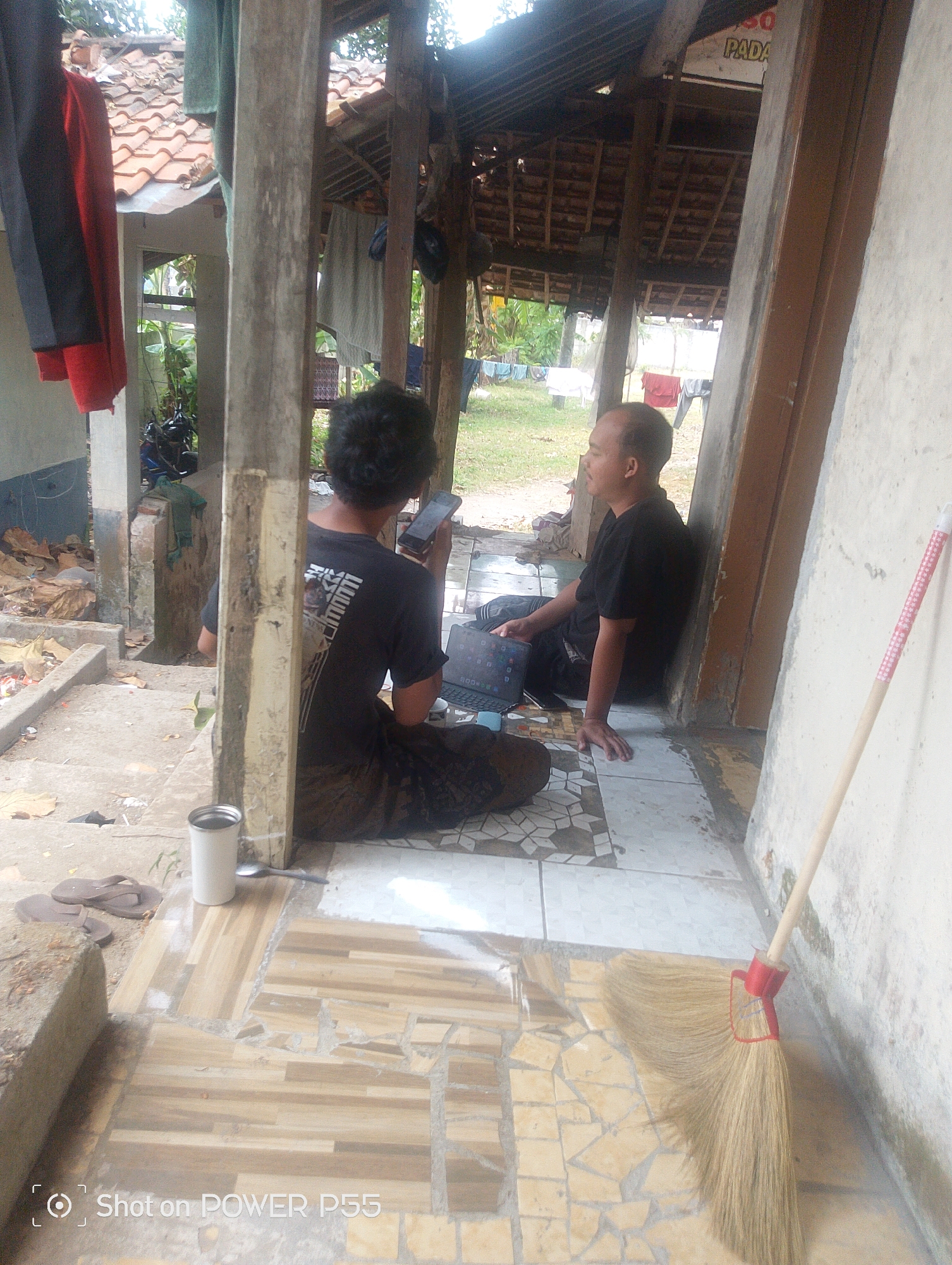 In Lebaksiu, Indonesia, two men are seen sitting on the ground in front of a simple, open-air structure. The man on the right, with a bald head and wearing a black t-shirt, is leaning against a wall and looking to his left. He has a laptop open in front of him. The man on the left, with dark, curly hair and wearing a black t-shirt and patterned pants, is facing away from the camera, holding a mobile phone in his right hand and looking at it. He also has a laptop in front of him. The ground is tiled in a mosaic pattern. To the left of the men, there is a concrete step with a white mug and a pair of flip-flops on it. A broom with a red handle is leaning against the wall on the right side of the image. The background reveals a tiled roof of the structure, some hanging clothes, and greenery. The overall scene depicts a casual setting, possibly a home or a workshop.