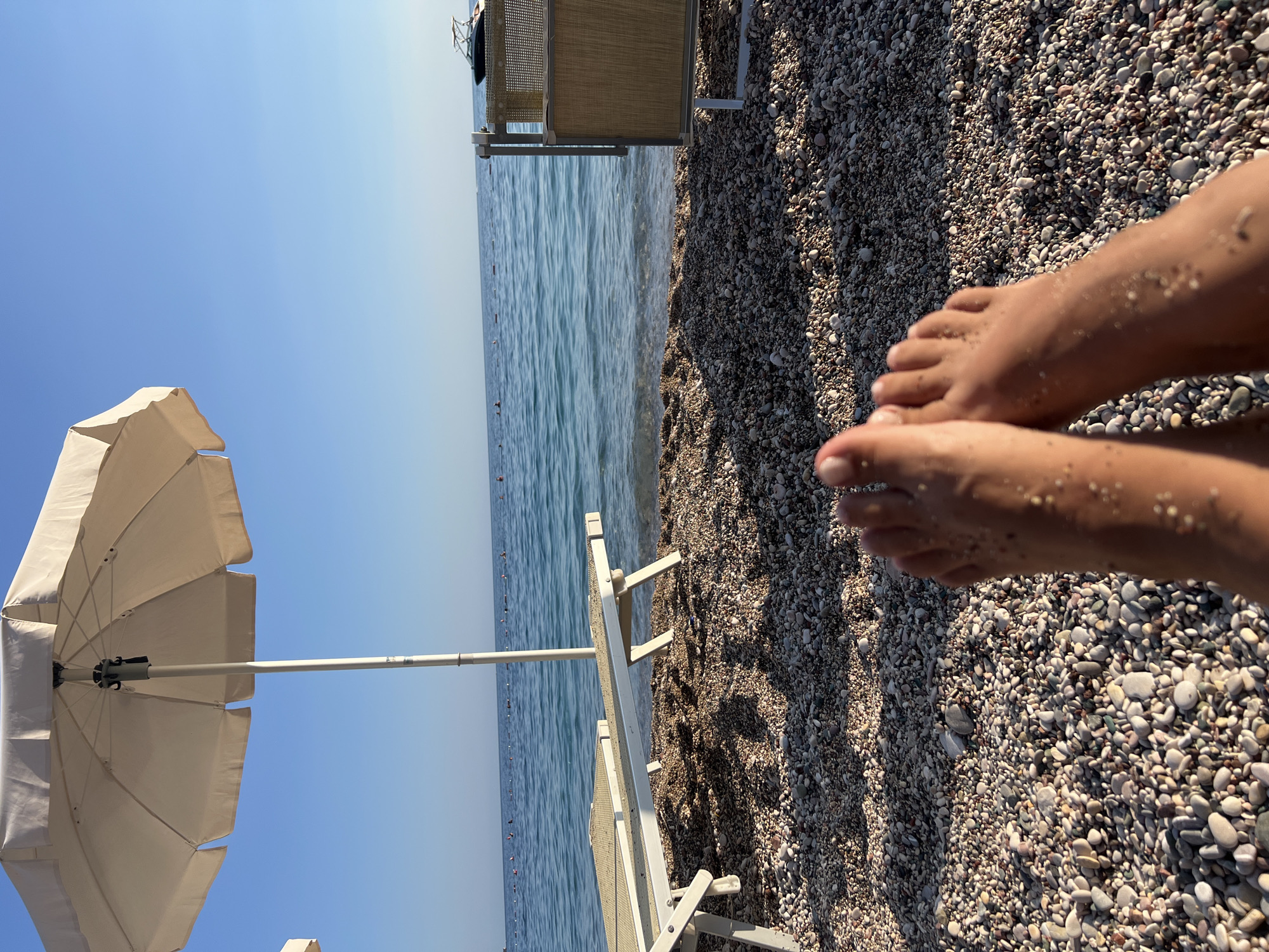 This media file captures a serene beach scene at Kamenovo, Montenegro. The foreground features a person's feet, lightly dusted with sand, resting on a textured shore of smooth pebbles. Beyond the feet, the calm, blue sea stretches to the horizon, its surface rippled by a gentle breeze. A white beach umbrella stands prominently on the left side of the frame, its wide canopy angled towards the sky. In the middle ground, a white sun lounger is visible, partially casting a shadow on the pebble beach. The clear blue sky dominates the upper portion of the image, suggesting a bright, sunny day. The overall atmosphere is one of relaxation and enjoyment of a summer day at the beach. No text is visible in the media file.