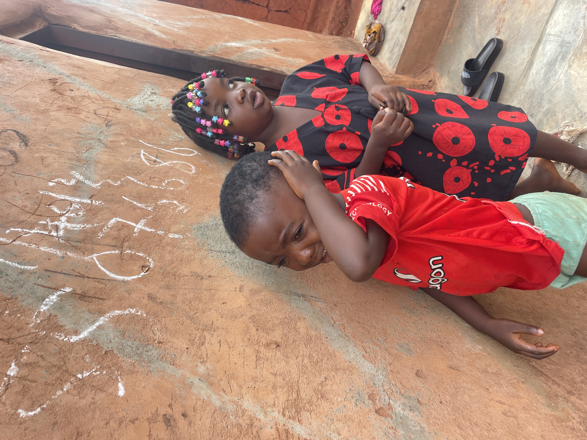 In Ogbe, Nigeria, two children are lying on a concrete surface near a reddish-brown wall with white markings. The child in the foreground is wearing a red shirt and light green shorts, and is holding their hands to their head. The other child is wearing a black dress with red circular patterns and has colorful beads in their hair. A pair of sandals is visible in the background, near the upper right corner of the frame.