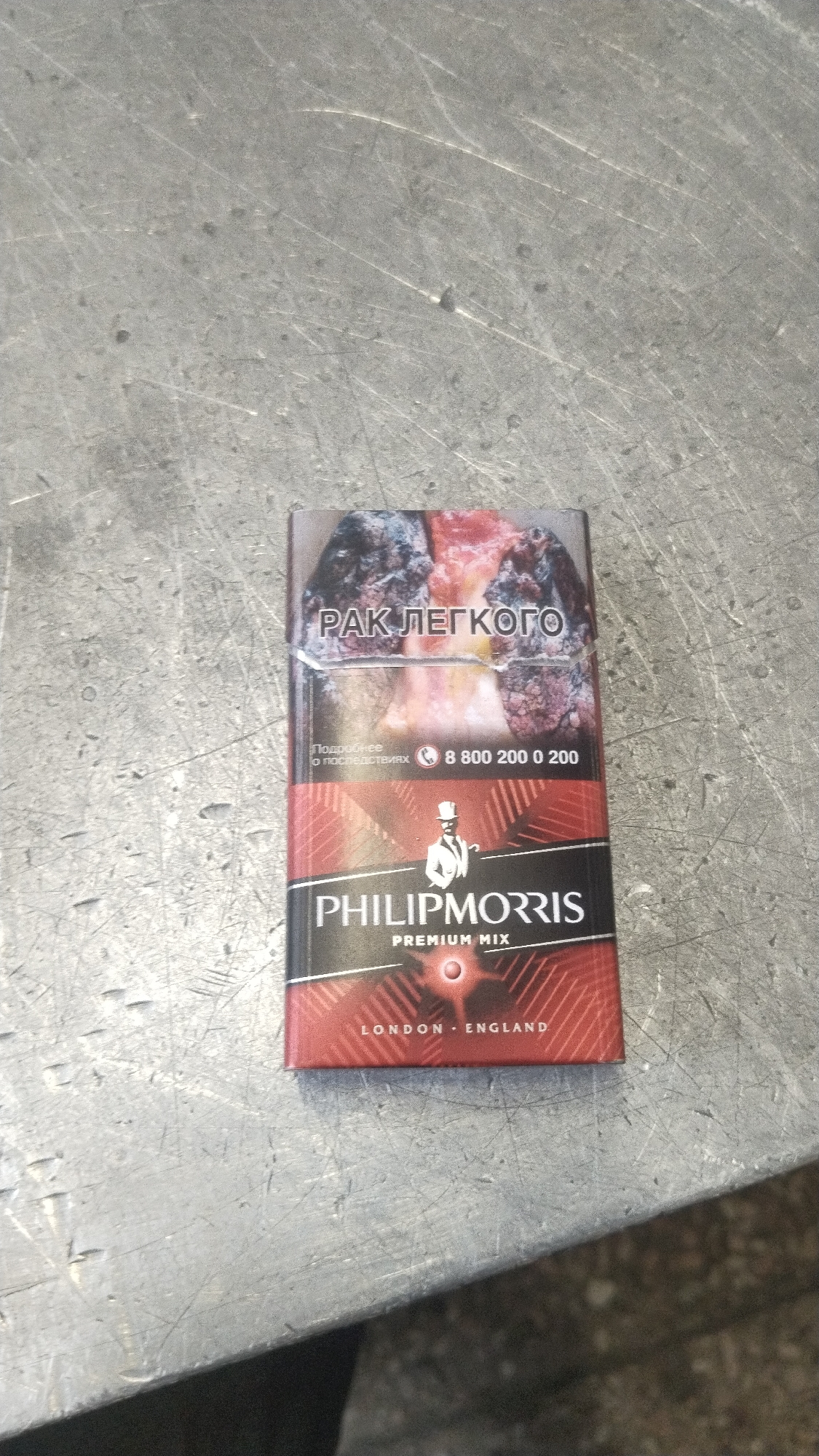 A close-up, top-down shot features a pack of Philip Morris Premium Mix cigarettes resting on a scratched, light gray, possibly metallic or concrete surface. The surface shows various scuffs, marks, and subtle indentations, suggesting a well-used tabletop or workbench. The cigarette pack is predominantly red with a black horizontal band across its middle.
The top portion of the pack displays a graphic health warning, characteristic of tobacco products. Visible text on the warning label includes "РАК ЛЕГКОГО" (LUNG CANCER) in white capital letters against a dark, possibly diseased lung image. Below this, smaller text reads "Подробнее о последствиях" (More about the consequences) and a Russian toll-free number "8 800 200 0 200" is also visible.
The black band on the pack features the brand name "PHILIP MORRIS" in white, below which "PREMIUM MIX" is written in a smaller font. A stylized figure in a top hat, holding a cane, is depicted above the brand name. Towards the bottom of the pack, "LONDON • ENGLAND" is printed, likely referring to the brand's heritage or design origin. No activity is taking place; the image is a static capture of the cigarette pack. The Russian text on the warning label is a strong indicator of the product's market.