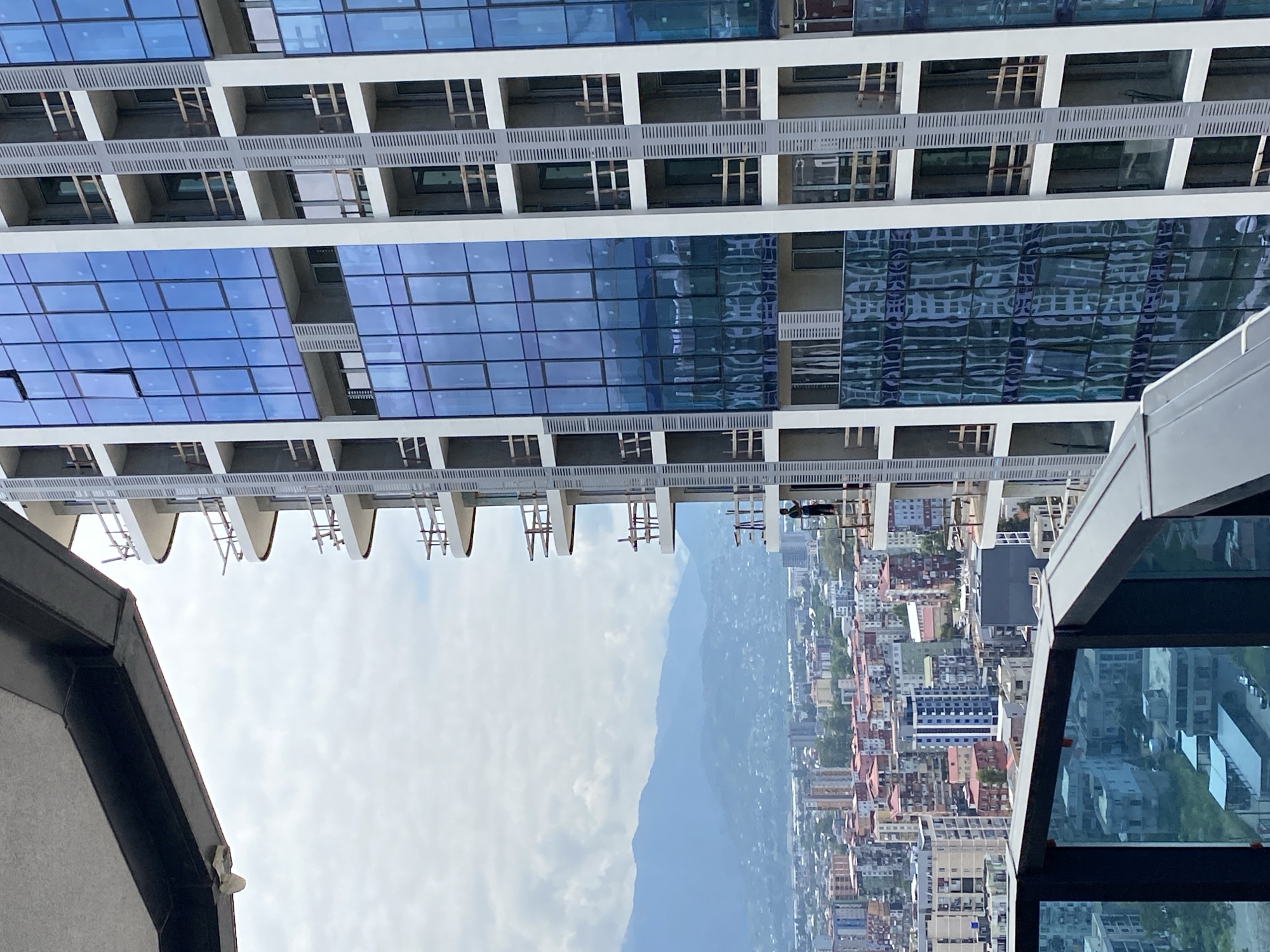 The image, taken from a high vantage point in Batumi, Georgia, showcases the ongoing construction of a modern high-rise building with a distinctive blue glass facade. The building features multiple balconies with scaffolding, suggesting active development. In the foreground, a portion of another building's balcony railing is visible, also overlooking the cityscape. A lone construction worker stands on a balcony of the new building, engaged in work. The background reveals a densely populated urban environment with various buildings, some completed and others under construction, stretching towards a range of mountains under a cloudy sky. The scene captures a moment in the dynamic development of Batumi's skyline.