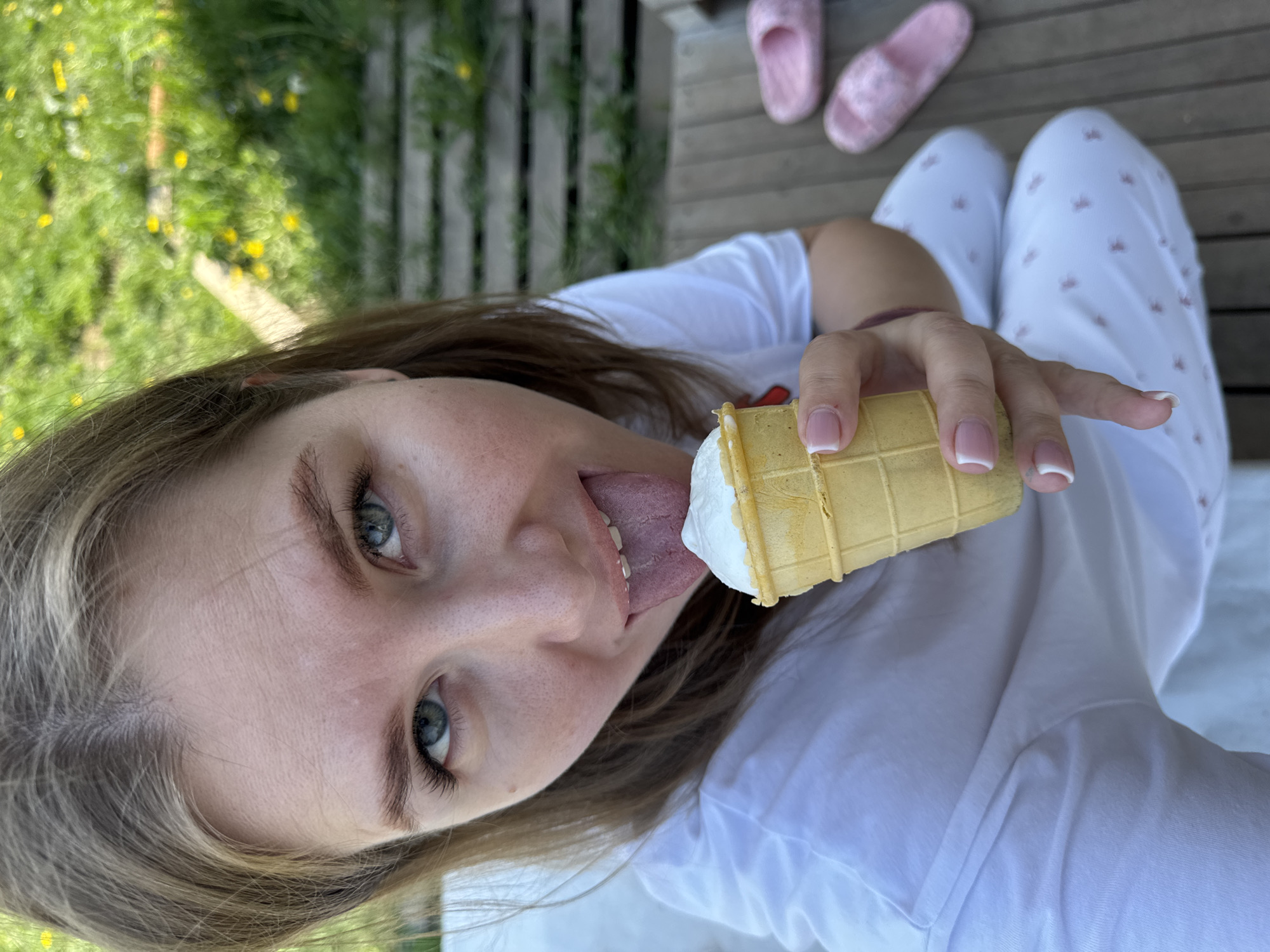 A fair-skinned young woman is captured in a close-up, upward-angled selfie while outdoors in the vicinity of Ферма, Russia. She is shown wearing a white t-shirt and white pajamas patterned with small red shapes. Her hair is light brown, and her eyes are blue. Her tongue is extended to lick an ice cream cone, which she holds in her right hand. The ice cream is white, and the cone is of a waffle design. Her fingernails are painted with a French manicure. In the background, a grassy area with yellow flowers and wooden decking are visible. A pair of pink slippers are also visible in the upper right corner of the image.
