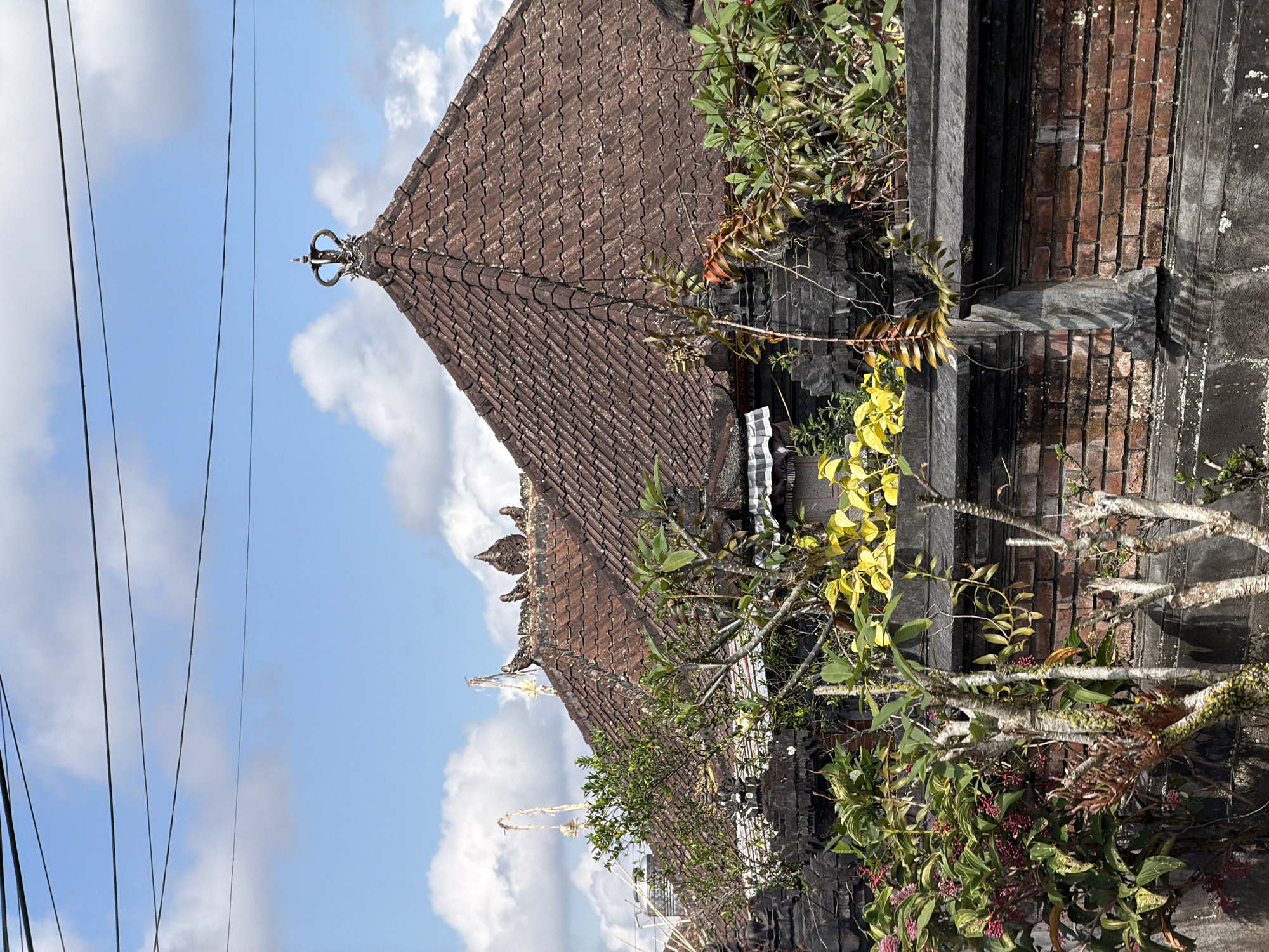 This image depicts the tiled roofs of traditional buildings in Baturiti, Indonesia, against a backdrop of a blue sky with scattered clouds. The roofs are made of reddish-brown, weathered tiles and are topped with decorative elements. On the rightmost roof, a prominent finial resembling a crown is visible. In the foreground, lush green and yellow foliage partially obscures the view of the buildings. A low wall constructed of brick and stone runs along the bottom of the frame, separating the viewer from the structures. Electrical wires are strung across the top of the image. The scene appears to be an exterior view of a residential or temple complex.