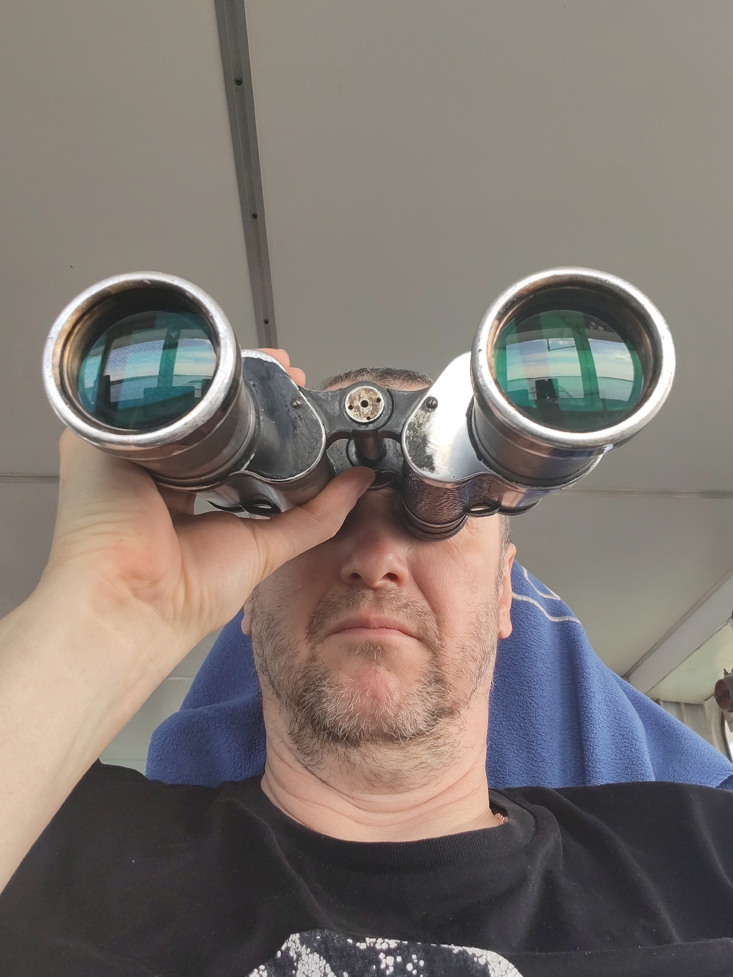 A man with a light beard and wearing a black t-shirt is holding a pair of large, silver binoculars up to his eyes. The lenses of the binoculars reflect a scene that appears to be outdoors, possibly a body of water. Behind the man is a blue towel and a white ceiling with a metal track. The location is unknown and not possible to determine from the image.