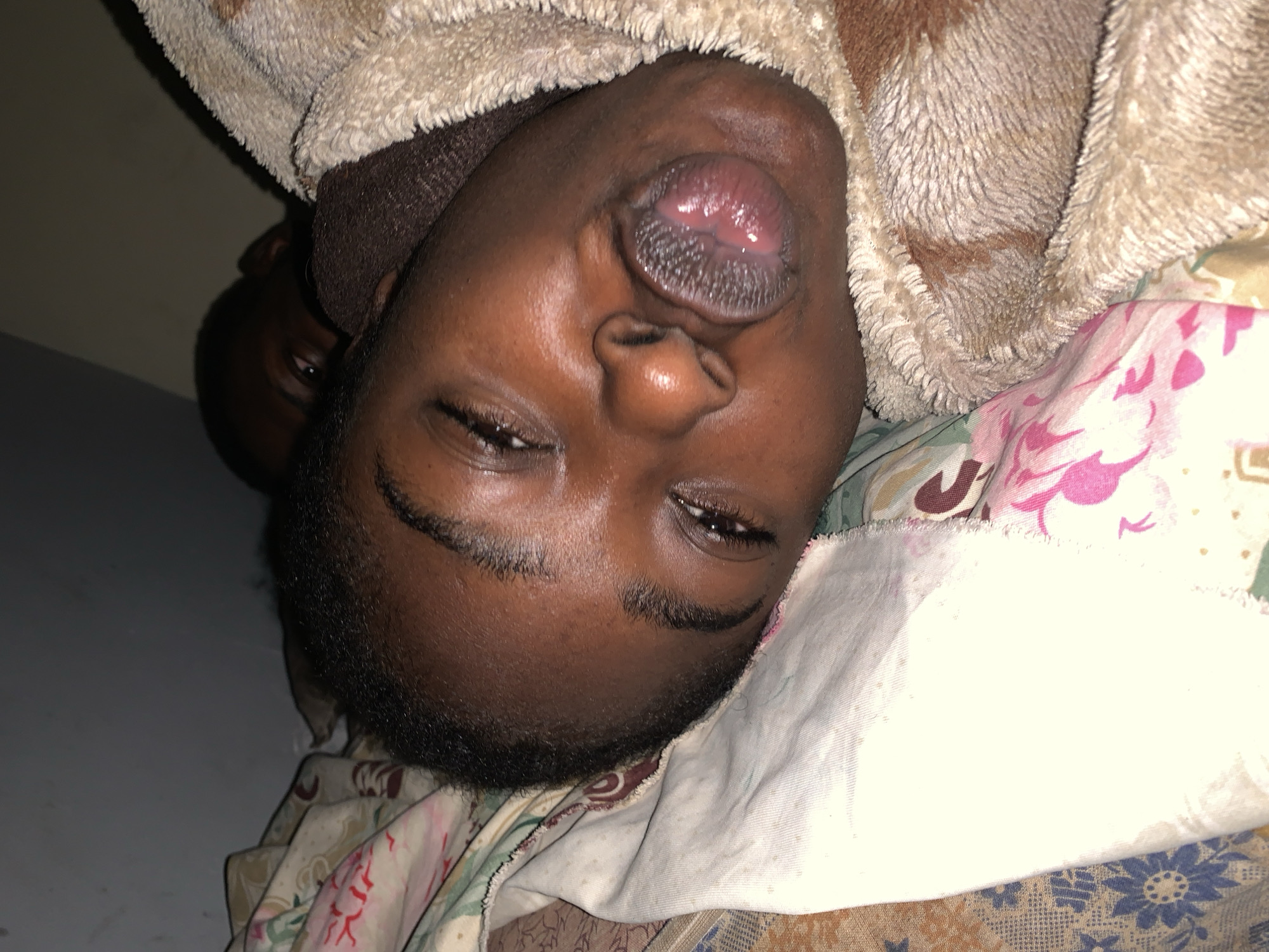 An eye-level, indoor shot in Rafin Par, Nigeria, shows a dark-skinned individual lying on a bed with floral patterned sheets and a white blanket. The person is upside down in the frame, their mouth slightly open. Part of a brown-and-beige textured blanket is around their head. A second person is partially visible in the background on the left side of the frame. The wall is grey.