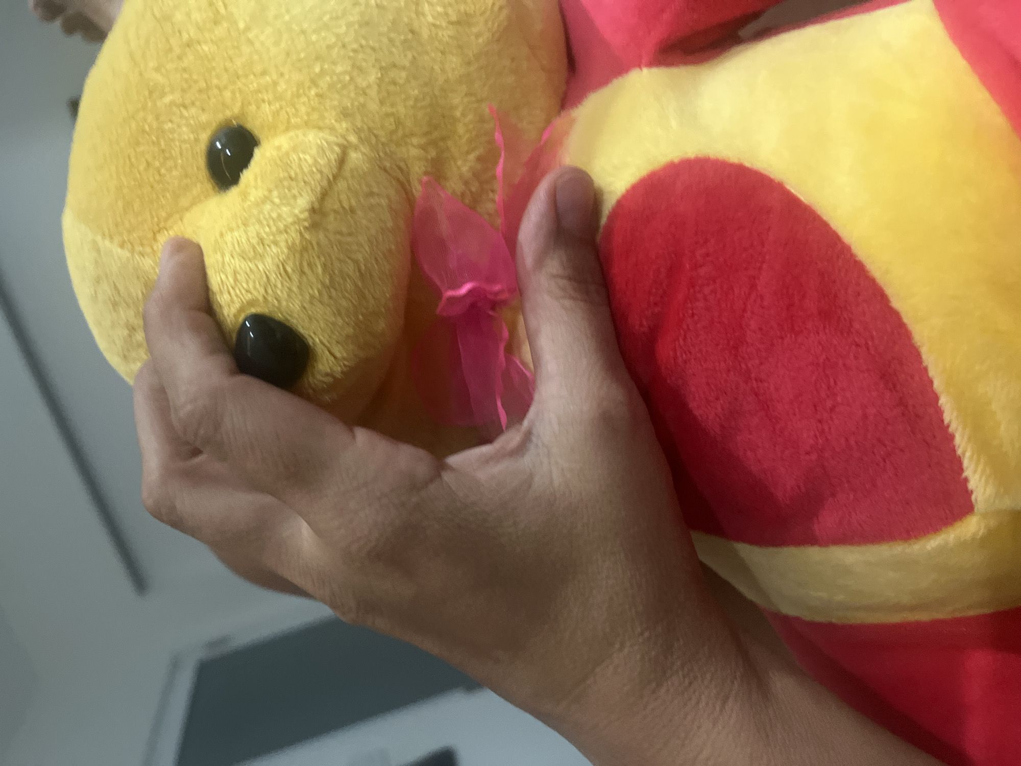 A close-up, chest-level shot shows a human hand holding a yellow stuffed bear. The hand, appearing to be that of an adult, is positioned on the bear's head, obscuring part of its face. The bear has two black button eyes and a small pink bow tied around its neck. The bear's chest is adorned with a large red heart shape. The background is softly blurred, suggesting an indoor setting with a gray wall and a hint of a doorway. No specific location in Nha Trang Ward, Vietnam, can be identified from the provided image.