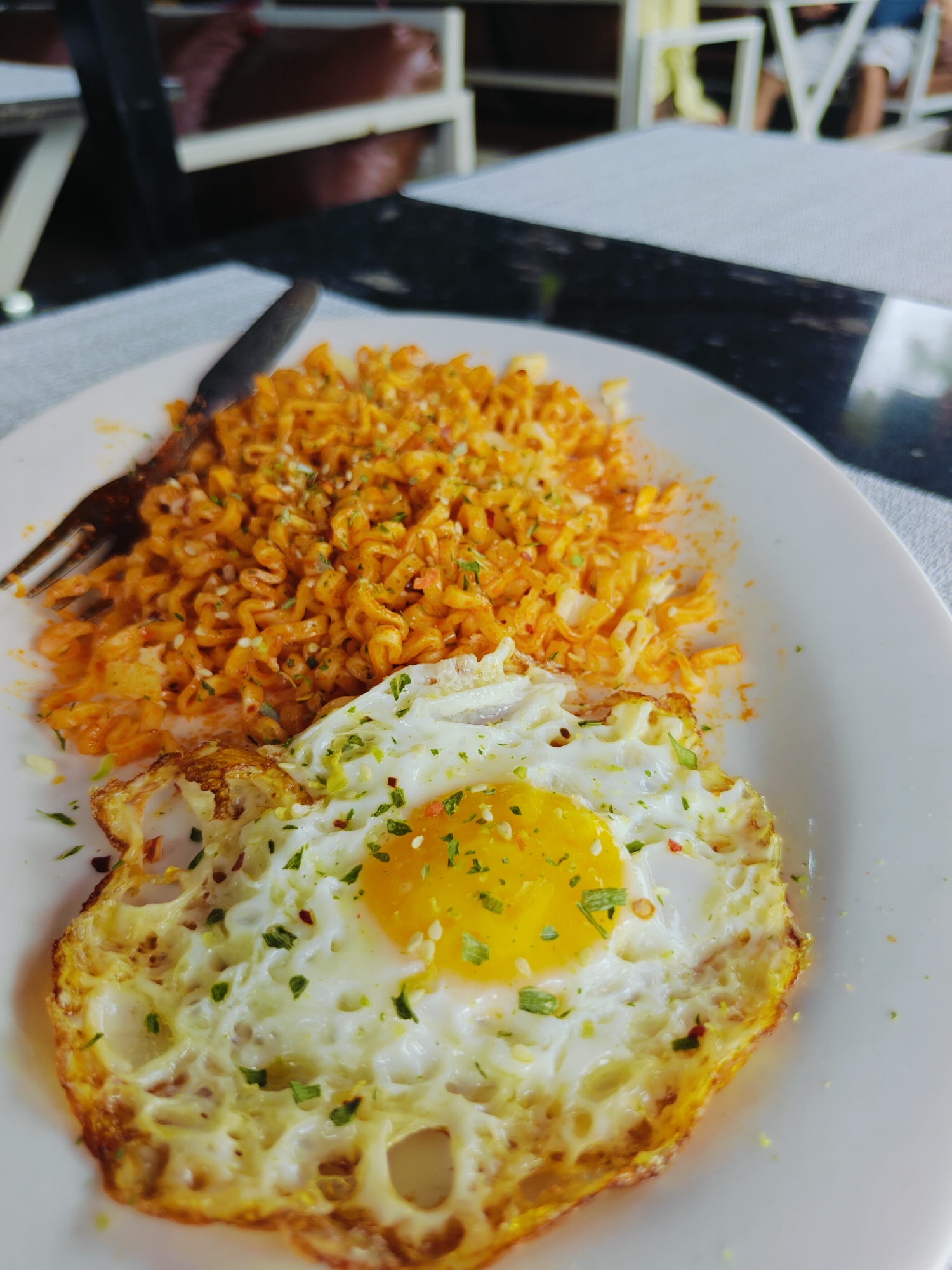 A close-up, eye-level shot shows a white plate of food with instant noodles and a fried egg, likely from a restaurant in Pokhara, Nepal. The noodles are a vibrant orange, coated in sauce and sprinkled with sesame seeds and herbs. A fork rests on the left side of the plate, partially submerged in the noodles. The fried egg is centered on the bottom half of the plate, with a bright yellow yolk and crispy, golden-brown edges, also garnished with herbs and red pepper flakes. The background is blurred, showing outdoor seating with tables and chairs, suggesting a casual dining setting. The lighting is natural, highlighting the textures and colors of the food.