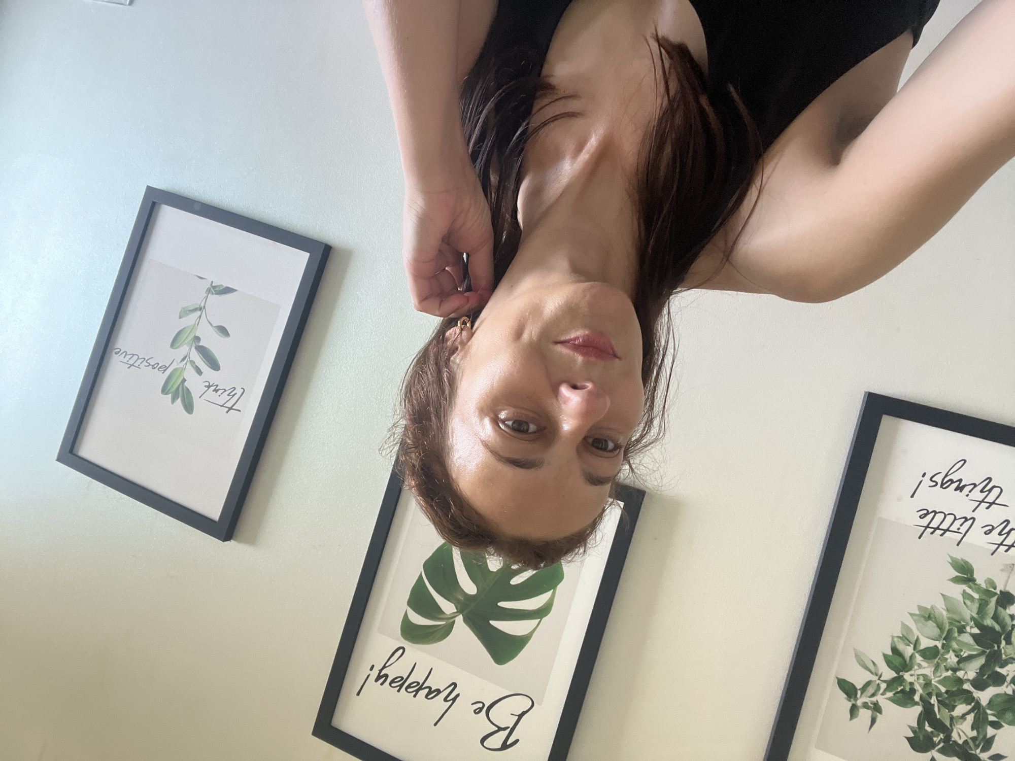 In a medium shot, a woman with shoulder-length brown hair, wearing a black sleeveless top, poses for a selfie against a light-colored wall. Her right hand is raised to her right ear, touching a small hoop earring. She is looking towards the camera with a slight smile, and a dimple is visible on her left cheek. Behind her, three framed pictures hang on the wall. The leftmost picture shows a leafy plant with the text "the little things!". The middle picture features a monstera leaf and the text "Be happy!". The rightmost picture displays a sprig of greenery with the text "think positive". The lighting suggests an indoor setting, possibly a home, with natural light illuminating the scene. The location is Unknown, Unknown.