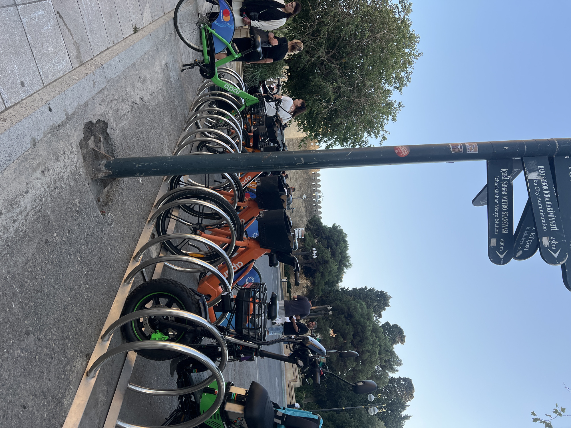 In Sabail Raion, Azerbaijan, a street signpost displays directional arrows and distances. The signpost is positioned near a rack of bicycles, some of which are green and orange. Several individuals are present, including women in casual attire and men standing near a road. The background features trees and a stone wall. The scene appears to be an urban environment with a public transportation service for bicycles.