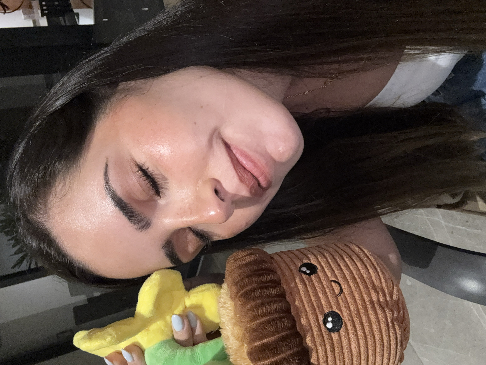 A woman with long, dark hair is lying down, holding a plush toy. The toy is brown with a cartoon face and yellow and green accents. She appears to be indoors, with dark furniture visible in the background. The specific location in San Bernardino Tlaxcalancingo, Mexico is not discernible from the image.