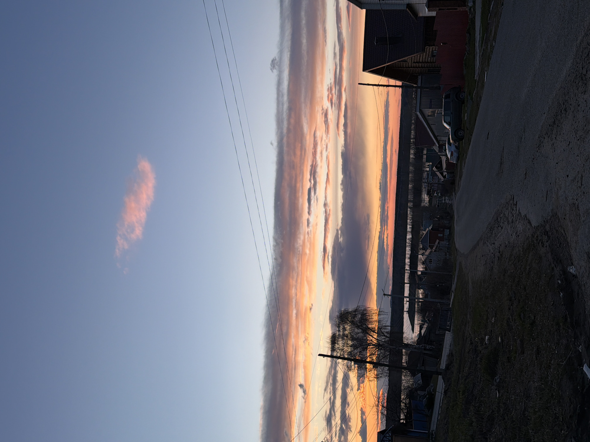 This image captures a serene street scene in Chaikovsky, Russia, at either sunrise or sunset. The sky is the dominant subject, featuring a magnificent display of clouds illuminated in warm hues. A prominent horizontal band of clouds stretches across the middle of the frame, glowing in shades of orange, pink, and yellow, indicative of the sun's low position. Above this, the sky is a clear, light blue, with a small, isolated reddish-orange cloud visible in the upper left.
In the foreground, a dirt or gravel road curves gently along the right side of the image, leading into the distance. To the left, several utility poles with multiple wires are visible, accompanied by silhouetted bare trees, suggesting late autumn or early spring. Scattered along the left side of the road are various modest houses or buildings, some partially obscured by the trees and poles. A dark-colored SUV and another car are parked near one of the houses. In the background, beyond the residential structures, a body of water, likely the Kama River, can be seen reflecting the sky's light.
The time of day is clearly sunset or sunrise, given the vibrant and warm colors of the sky and the overall lighting. The weather appears calm and clear, with distinct cloud formations. No people are visible, and no particular activity is taking place, contributing to the quiet, peaceful ambiance of the scene. There is no visible text in the image.