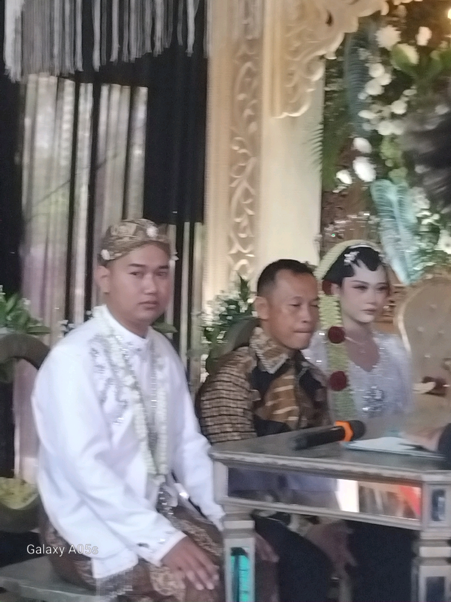 In an indoor setting, likely a wedding ceremony in Karanganyung, Indonesia, three individuals are observed. On the left, a man wearing a traditional white ceremonial shirt with intricate silver embroidery and a patterned headpiece sits, facing slightly to the right. To his right, a man in a patterned brown and black shirt is seated and looking to his right. Further to the right, a woman in a white wedding gown and a traditional Javanese headdress, adorned with flowers, is also seated and looking to her right. A reflective table is positioned in front of the seated individuals, with a microphone and some papers on its surface. The background features decorative fabric drapes and floral arrangements, suggesting a festive occasion. The "Galaxy A05s" watermark is visible in the lower left corner of the image.