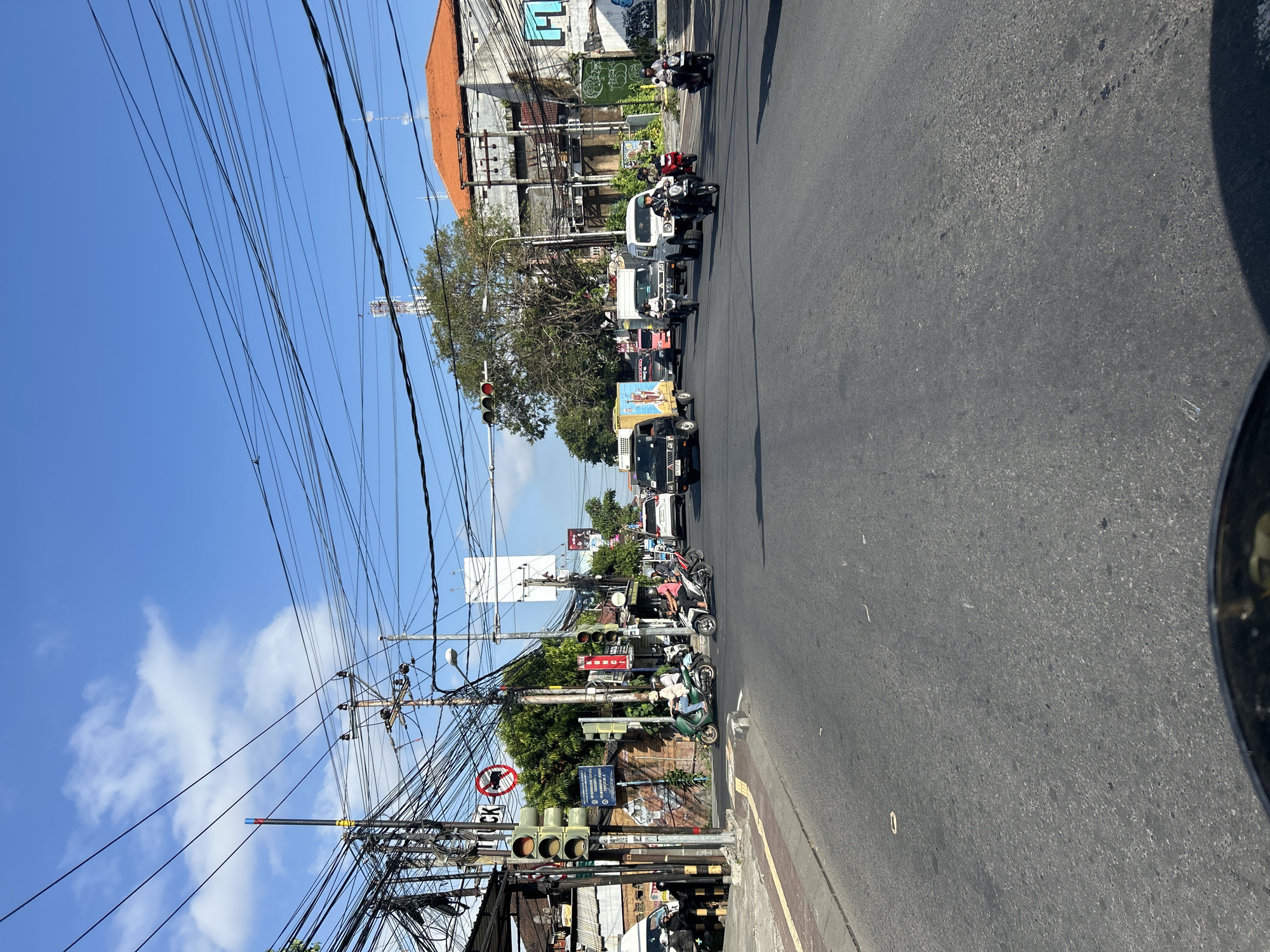 This image depicts a street scene in Denpasar, Indonesia, on a bright, sunny day with a clear blue sky dotted with white clouds. The road is paved and appears to be a busy intersection, with numerous vehicles and motorcycles.
In the foreground, the perspective suggests the photo is taken from a vehicle, possibly a motorcycle, as a curved black edge, likely a part of the vehicle's body or mirror, is visible at the bottom. The road itself has lane markings and some debris.
On the left side of the road, there's a sidewalk with a brick border. A traffic light pole with three signal lights, currently red, stands prominently. Next to it, a pole with a circular sign featuring a red border and a diagonal line over a motorcycle symbol indicates "no motorcycles." There are also various signs and graffiti on a wall behind the sidewalk. Several motorcycles and a white van are parked near the intersection.
The center and right of the image are filled with traffic. A variety of vehicles, including cars, vans, trucks, and numerous motorcycles, are moving through the intersection. Some of the motorcycles are carrying one or two riders, many wearing helmets. A prominent orange-roofed building is visible on the right side, with other structures and trees in the background. A substantial amount of overhead electrical wiring is a dominant feature, crisscrossing the sky, typical of many urban areas in Indonesia. The overall impression is a bustling urban environment with active transportation.