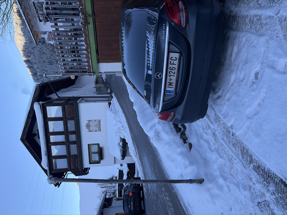 The scene depicts a winter landscape in Sankt Jakob am Arlberg, Austria, characterized by snow-covered terrain under a clear, bright sky. In the immediate foreground, the rear of a dark grey Mercedes-Benz C300 sedan is parked on snow, displaying an Austrian license plate "IM • 126 FC". Tire tracks are visible in the snow on the ground around the vehicle.
To the left, a rustic wooden pole stands upright beside a paved road that inclines uphill and curves towards the left. Snow banks line the edges of the road. Further up the road on the left, a dark-colored station wagon, possibly a Skoda, is partially visible.
The central feature is a traditional chalet-style building with a white rendered facade and dark brown wooden elements. It features a prominent enclosed balcony or bay window with multiple panes on its upper level, covered with a layer of snow. Below this, on the ground floor, is a smaller window with a visible flower box beneath it, and an ornate, decorative metal gate motif affixed to the wall. Snow is piled against the base of this building.
To the right of this building, partially obscured by the Mercedes, is another structure with a dark wooden garage door and a slatted wooden fence above it. Smoke or vapor is observed rising from a metal chimney pipe near this area.
The background features snow-covered mountains densely forested with coniferous trees, illuminated by direct sunlight on their upper slopes. No individuals are present in the image.