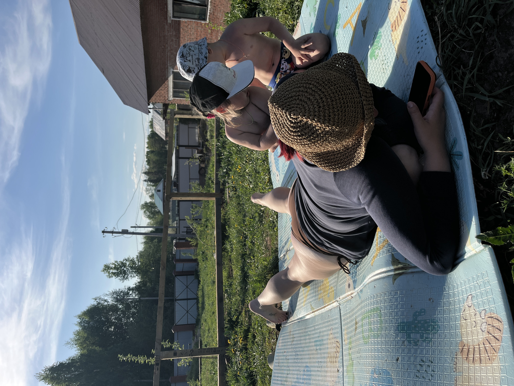 The image depicts three individuals outdoors on a hot day. A woman in a dark dress and a straw hat is seated on a blue and white textured surface, possibly a plastic play pool or mat. She is looking at a smartphone in her right hand. Two children, one wearing a patterned bucket hat and swimwear and the other wearing a black and white baseball cap, are seated near her on the same surface. They appear to be playing or interacting with each other. The background includes a grassy field with wildflowers, a wooden fence, trees, power lines, and a building with a brick exterior. The location is identifiable as being near the city of Chaikovsky, Russia due to the architectural styles and common Russian residential areas.