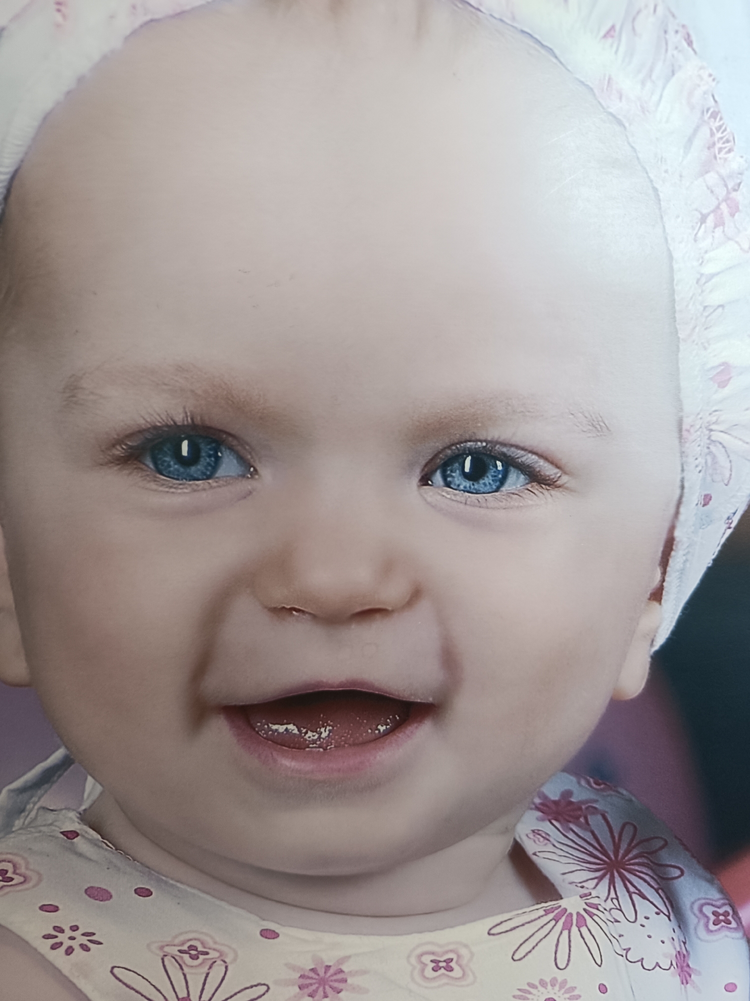 A close-up shot depicts a baby with fair skin, bright blue eyes, and a joyful expression. The baby is wearing a floral-patterned dress and a matching bonnet. The baby's mouth is open, revealing a glimpse of its tongue. The photograph's location in Pleven, Bulgaria, is unknown.