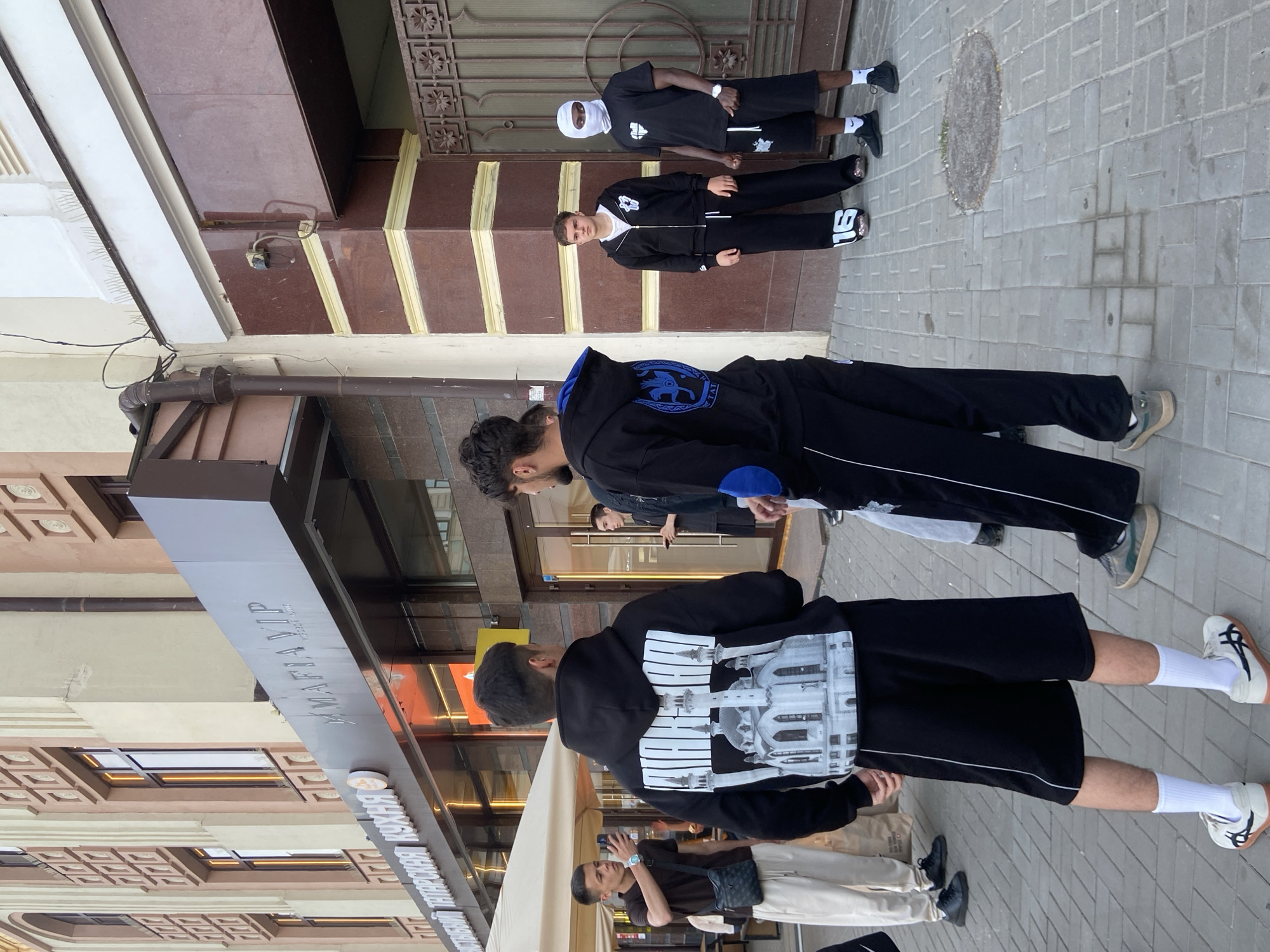 The image shows a group of people on a sidewalk in Kazan, Russia. The group includes at least six individuals of varying heights, ages, and ethnicities. They are dressed in casual clothing, with several wearing athletic-style tracksuits or hoodies with designs. One person wears a white head covering. The people are standing near a building with a sign that reads "MAFIA VIP" in Cyrillic script. The building appears to be a restaurant or entertainment venue. The surrounding environment includes a paved sidewalk, a street, and other buildings. The lighting indicates that the photograph was taken during the day.