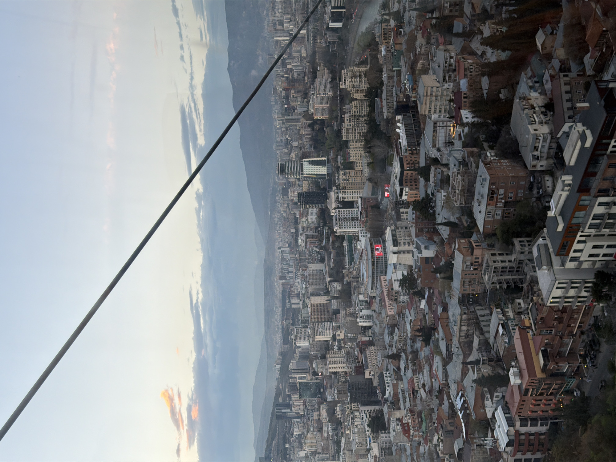 The image shows an aerial view of the city of Tbilisi, Georgia, likely taken from a cable car. The foreground features a cable wire running diagonally from the top left to the bottom center of the frame. In the background, a cityscape stretches out, densely packed with buildings of varying heights and architectural styles. A mountain range is visible beyond the city, partially obscured by clouds and haze. The sky transitions from a light, clear hue on the left to a more overcast tone near the mountains. The urban landscape is a mix of residential and commercial structures.